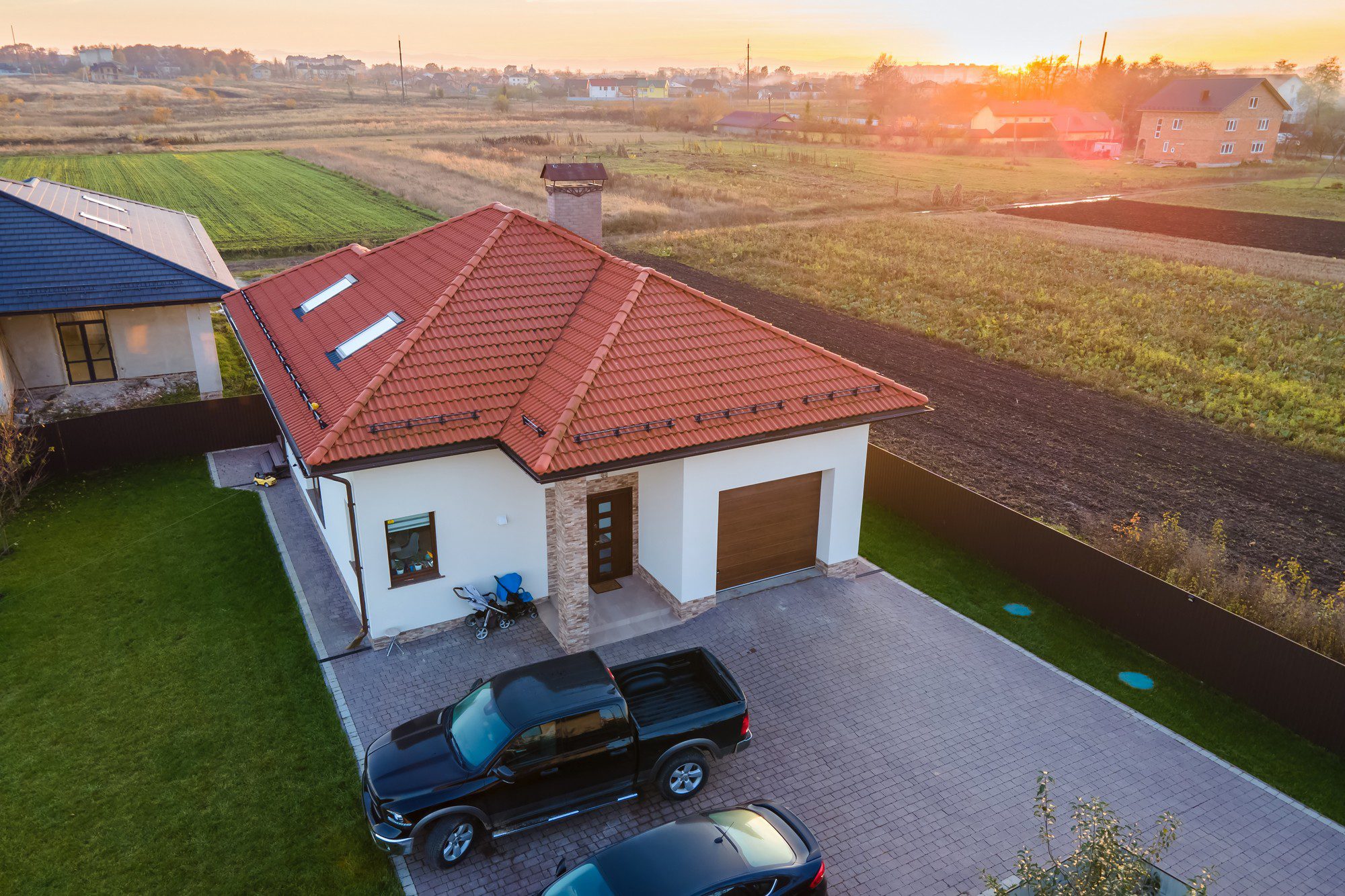 This image shows an aerial view of a residential property during what seems to be sunrise or sunset, as indicated by the warm hues in the sky. The house has a red tiled roof and is surrounded by a manicured green lawn. There's a paved driveway with two vehicles parked outside—one SUV and a sedan. Adjacent to the house, there's a structure with a dark roof, possibly an older building or a barn. The surrounding landscape includes open fields, and in the background, there are more residential buildings. A fence borders the property on at least two sides. The entire scene suggests a suburban or rural setting.