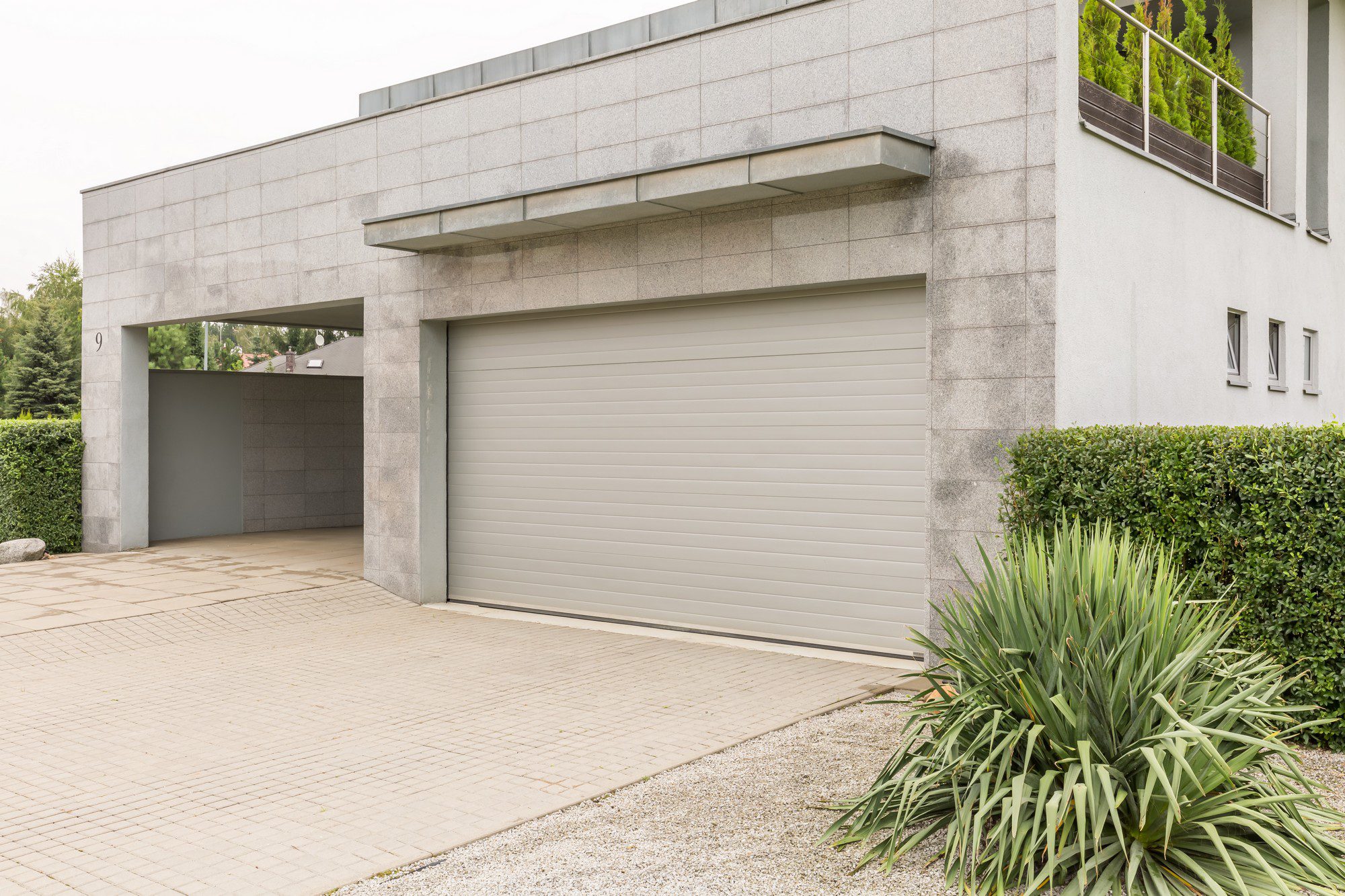 The image depicts a well-maintained garage attached to a large, modern house. The garage sports a sleek and modern design with a white sectional garage door that contrasts with the dark grey exterior walls of the house. The driveway leading up to the garage is expansive, paved with smooth, neatly arranged grey bricks, and is impeccably clean. Well-manicured shrubs and greenery border the driveway, adding a touch of nature to the contemporary setting. Large windows on the house offer glimpses of the interior, indicating a bright and spacious layout inside. The surrounding area is tidy, with a clear sky overhead contributing to the overall serene and upscale appearance of the property.