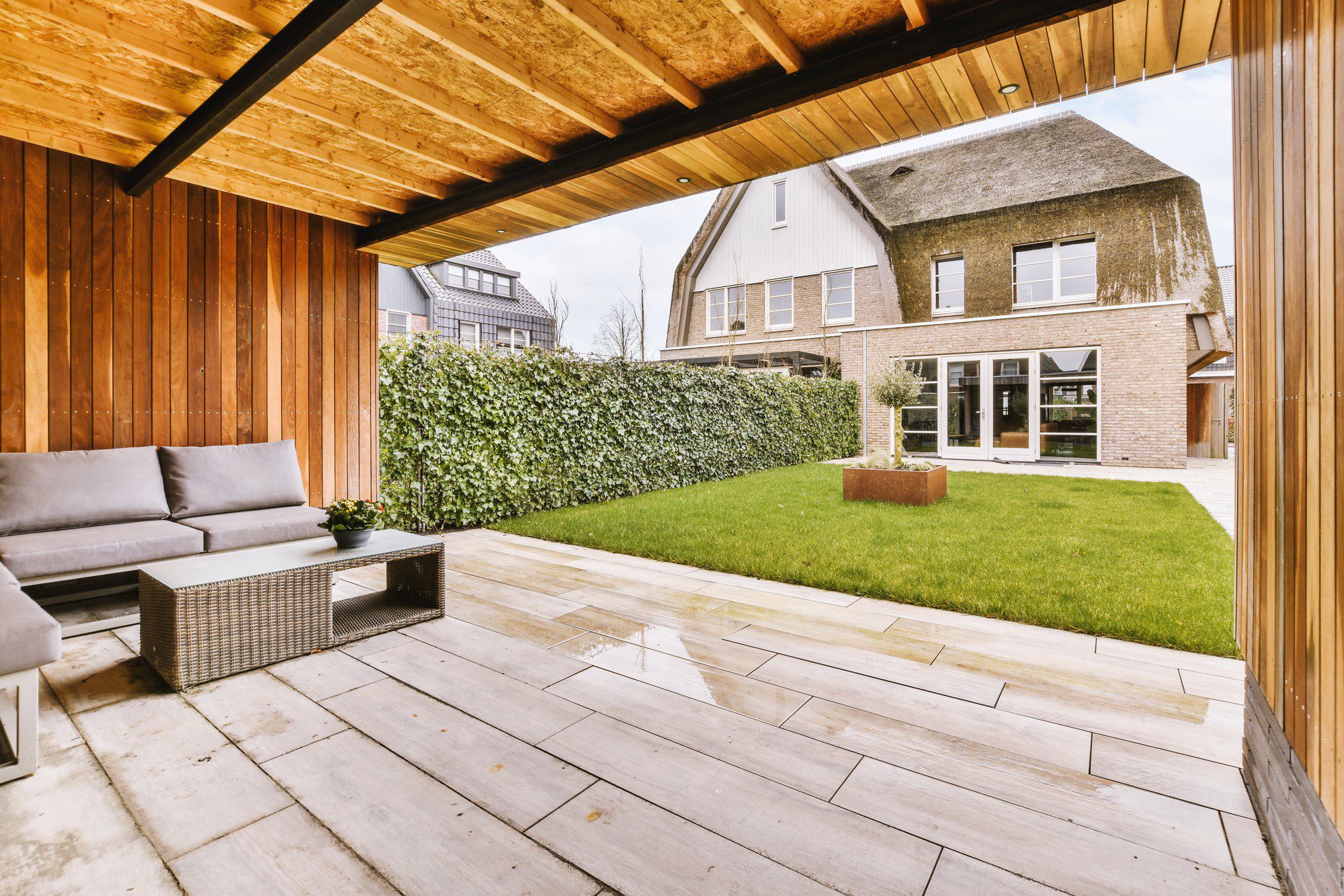 The image shows a modern backyard with a well-maintained garden space. Here are the key features visible in the image:1. A covered patio area with a wooden ceiling and beams that create a cosy outdoor space.
2. An outdoor lounge set consisting of a sofa with cushions and a matching wicker coffee table, suitable for relaxation and social gatherings.
3. A neatly trimmed green lawn that serves as the central garden area.
4. A wooden fence or wall on one side that provides privacy and enhances the aesthetic appeal with its vertical wood planks.
5. Paving stones that create a path and define the patio area, showing some water puddles which suggest recent rain.
6. A hedge that acts as a natural boundary on the other side of the garden, offering a lush green backdrop.
7. A large, modern house with big glass windows and doors, allowing plenty of natural light inside and ensuring a view of the garden from indoors.
8. A smaller raised bed or planter box with what appears to be small shrubs or plants, adding a touch of landscaping to the setting.The overall appearance is of a well-designed, contemporary outdoor living space that blends comfort with nature.