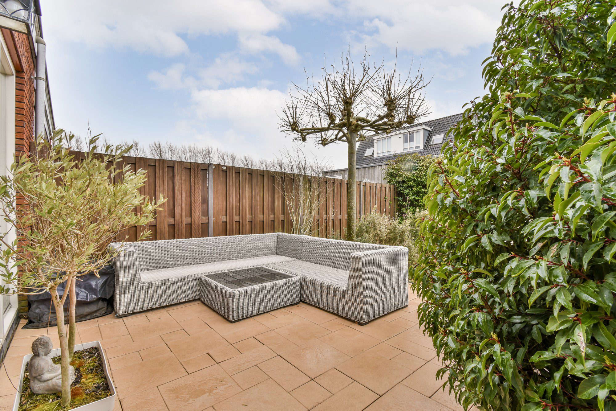 The image shows a neatly maintained outdoor patio area. There is a modern wicker furniture set comprising of a corner sofa and a square coffee table placed on tiled flooring. A young olive tree is potted and stands slightly to the left in the image, with a small stone buddha statue at its base. To the right, there is a thick, leafy green shrub. In the background, there's a wooden fence that provides privacy and some deciduous trees without leaves, which might suggest that the season is either autumn or winter. There are houses visible in the distance, indicating this patio is part of a residential area. The sky is mostly cloudy, implying an overcast weather condition.