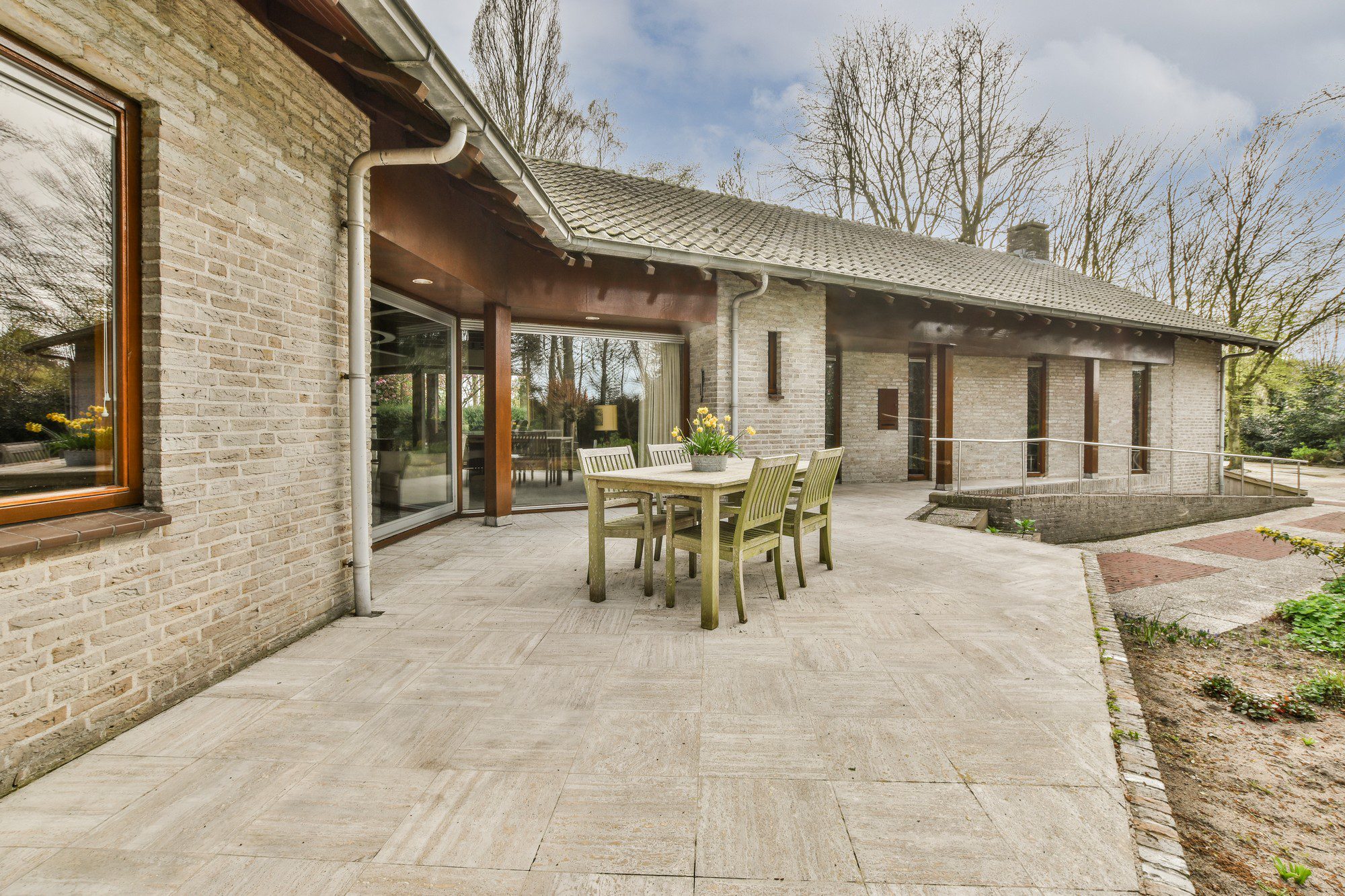 This is an image showing the exterior of a house with a tiled patio area. The house has light brick walls and a tiled roof. A dining set consisting of a wooden table and chairs is situated on the patio, indicating a space for outdoor dining or relaxation. Large glass windows or doors line the wall of the home facing the patio, offering a view inside the house and likely allowing natural light to enter. Around the patio, there's some landscaping visible, and trees can be seen in the background, suggesting the house is in a residential area with some greenery. Overall, the setting appears tranquil and comfortable for outdoor living.