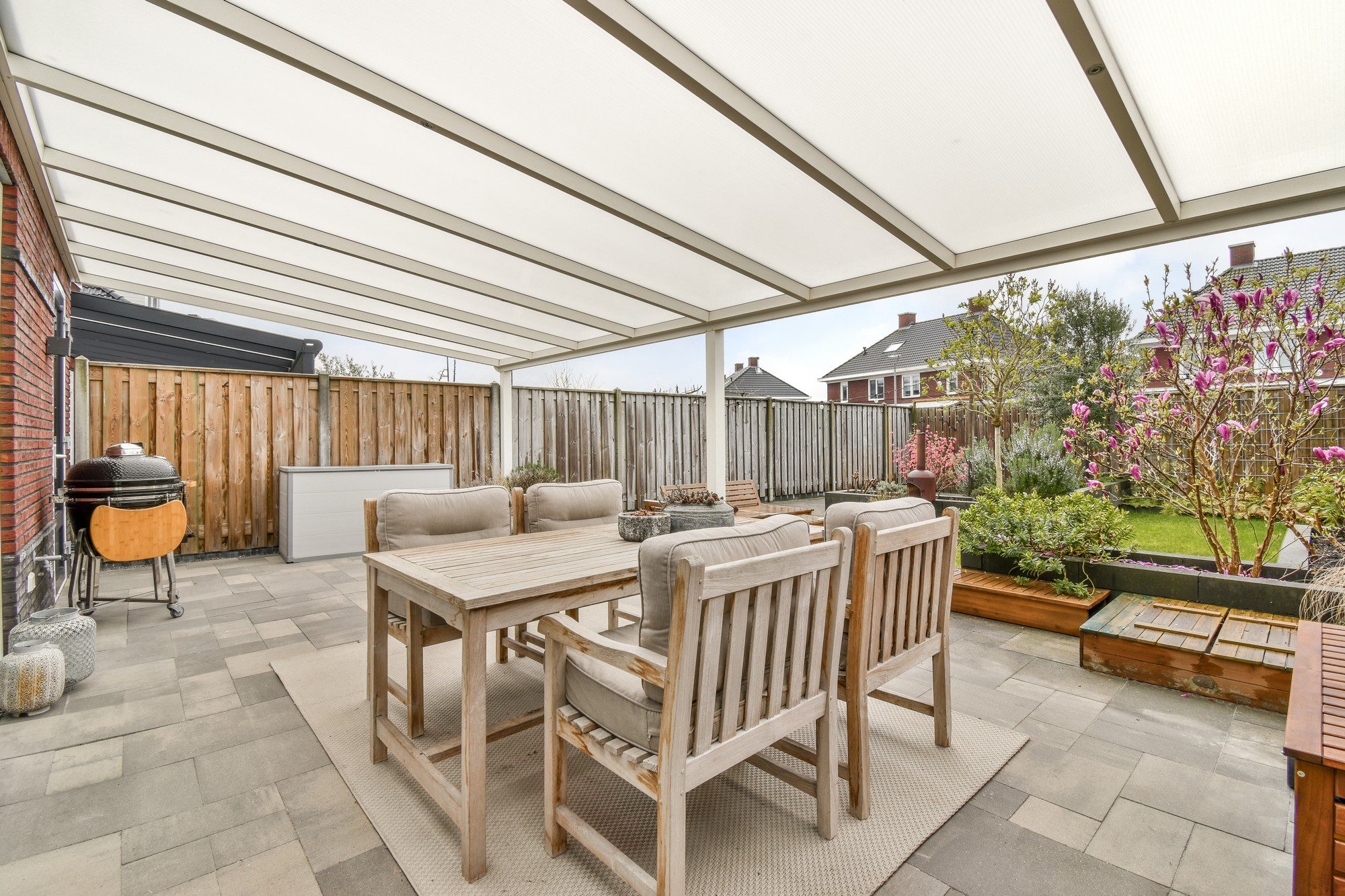 This is an image of an outdoor patio area. The space is covered by a white pergola, which provides shelter while still allowing some light to filter through. The patio is furnished with a wooden dining table set that includes a bench and chairs with grey cushions, creating a cosy outdoor dining space. There is also an outdoor kitchen or barbecue area on the left side, featuring a grill. Woven and patterned outdoor rugs are placed on the tile flooring to add warmth and texture to the setting.In the background, there is a wooden fence that encloses the backyard, providing privacy. Beyond the patio area is a well-maintained garden with green grass, a variety of plants, and some blooming pink flowers. The surrounding environment includes residential houses, suggesting the image is taken in a suburban neighborhood.