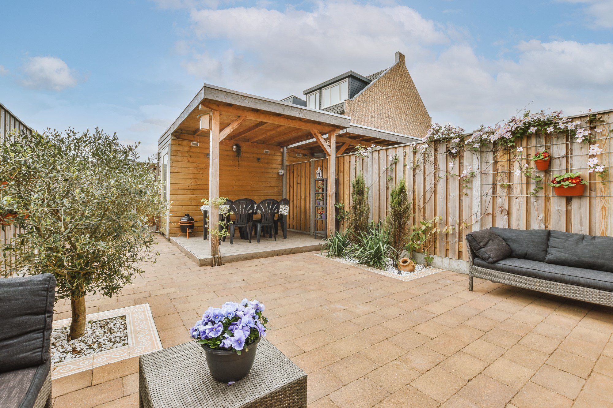 This image shows a cosy backyard patio area. The patio is paved with square stone tiles and surrounded by a wooden fence. On the left, there is an olive tree planted in a square section bordered by white stones. A wicker two-seater sofa with a thick black cushion is in the foreground on the right, with a small wicker table holding a pot of purple flowers in front of it.Centrally, there is a covered wooden structure that appears to be a gazebo or pergola with an open front, featuring a large wooden dining table with several chairs around it, providing a space for outdoor dining or gatherings. The back wall of this structure has shelves with various small decorative items, and it looks like a cosy, sheltered spot for relaxation.The wooden fence is adorned with climbing plants and flower pots, adding touches of greenery and colour to the space. There are a few other pots and decorations scattered around, which give the patio a lived-in and personal feel. The sky is partly cloudy, suggesting it might be a pleasant but not overly sunny day, perfect for enjoying time outdoors.
