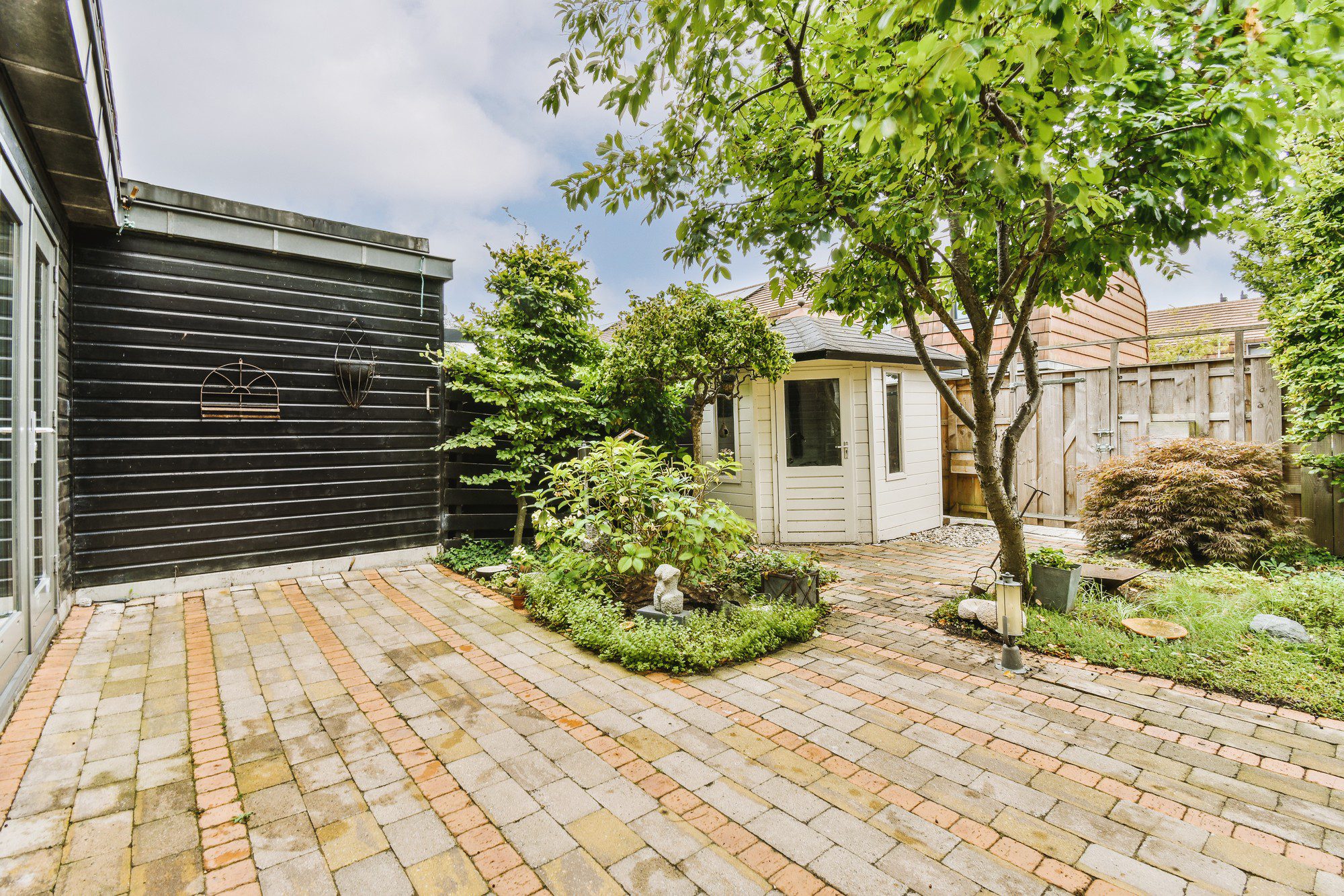 This image shows a cosy backyard patio and garden area. The patio is paved with red brick pavers arranged in a pattern, and there's a lush tree in the centre providing shade. The right side features a small, cream-coloured building that could be a shed or storage space, and there is a wooden fence at the back indicating the boundary of the property. On the left, a dark-painted exterior wall of a house is visible with two decorative metal pieces that resemble window frames mounted on the wall. The garden around the patio is well-maintained with an array of green plants and shrubs. Some garden decorations, like a bird bath and what appears to be a small statue, can be seen among the plants. The sky is partly cloudy, suggesting a temperate outdoor setting.