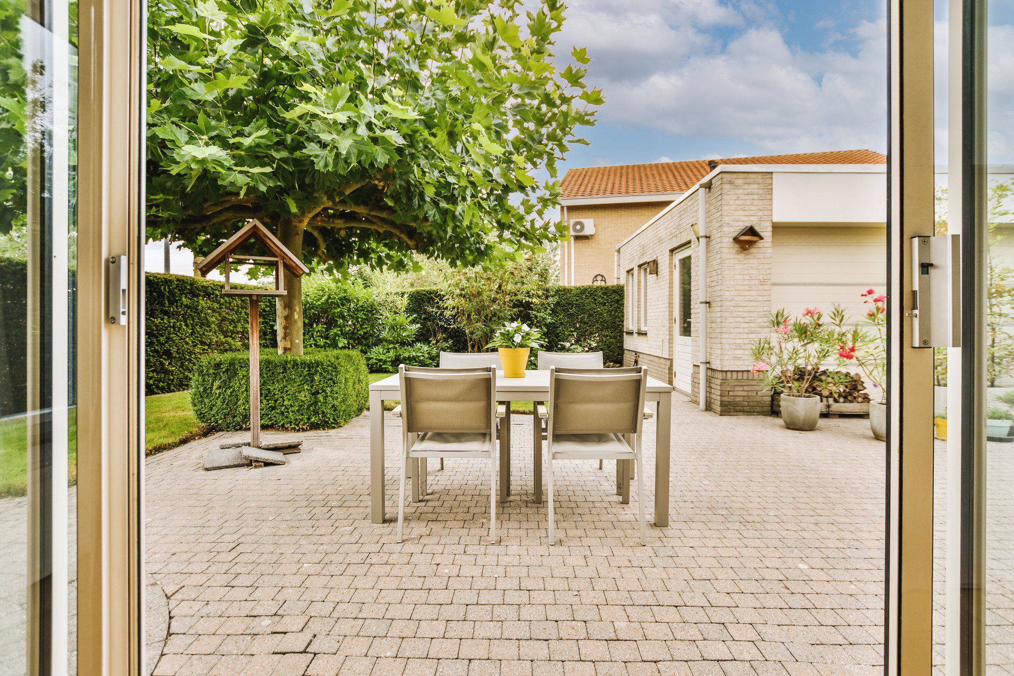 The image shows a patio area of a residential home. In the centre, there is an outdoor dining table with four chairs around it, set on paving stones. On the table is a potted plant in a yellow container, adding a touch of colour to the scene. Above the table is a large leafy tree, providing shade and greenery. To the left of the table, there is a birdhouse on a stand, and behind that, a neatly trimmed hedge.In the background, you can see the exterior of a house with a brick facade, windows, and a small section with lighter wall cladding or paint. There is a security camera mounted on the wall. In front of the house, there are more potted plants, some of which are flowering, enhancing the garden's aesthetic. The view is framed by an open glass door on the left, suggesting that the photo was taken from inside the house looking out onto the patio. The sky is partly cloudy, indicating a day with a mix of sun and clouds.
