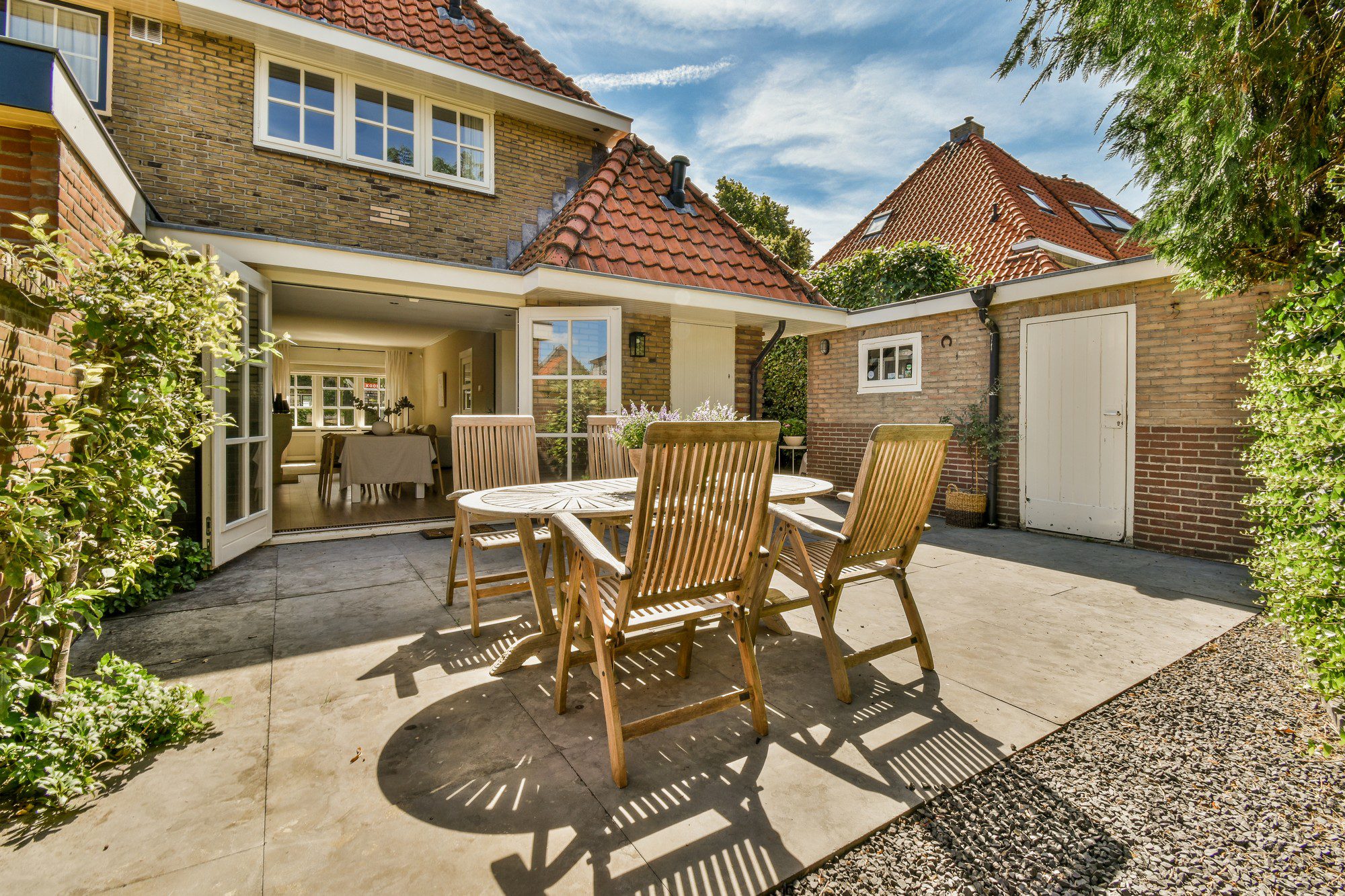 The image shows a sunny outdoor patio area of a house. There is a set of wooden garden furniture consisting of a round table and four folding chairs. The ground has a combination of paving stones and some areas with small pebbles.In the background, you can see the exterior of the house which has a brick facade and tiled roof, indicating a traditional architectural style. The house features double doors that open onto the patio, and above the doors, there is a canopy providing shade. Additionally, there is a white door to what may be a shed or storage area on the right side of the image.The scene is framed by various plants and shrubbery, hinting at a well-maintained garden space. The sky is blue with some scattered clouds, suggesting pleasant weather conditions. It looks like a calm and inviting space, ideal for outdoor dining or relaxation.