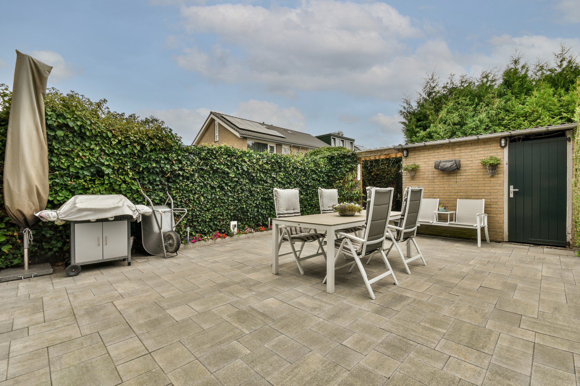 This image shows a neatly organized backyard or patio area. It features a large, paved surface with rectangular tiles. On the left side, there's a covered barbecue grill and a wheelbarrow against a hedge that borders the yard, giving a sense of privacy. There's an outdoor dining table set up in the centre with six chairs around it, all of which appear to be made of a light-coloured material that could be metal or plastic, with cushions on the seats for comfort. On the table is a small decorative plant. To the right, there's a brick wall with wall-mounted planters containing greenery and a green door, likely leading to a storage shed or another section of the property. The sky is a mix of clouds and blue, suggesting an overcast but pleasant day. Overall, the space looks well-maintained and inviting for outdoor dining or relaxation.