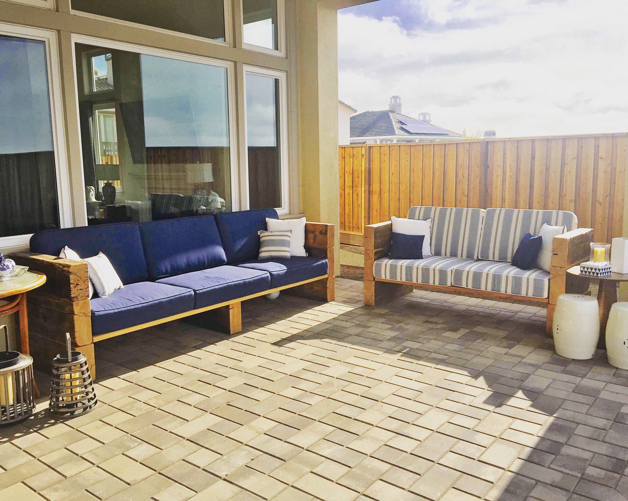 The image shows a nicely appointed outdoor patio area. There is a large sectional sofa with blue upholstery on the left and a smaller love seat with blue and white striped cushions on the right, both providing comfortable seating options. The flooring consists of a patterned arrangement of pavers. A wooden fence encloses the area, providing privacy.Decoration and additional furnishings include assorted pillows on the seats, a small table with what appears to be a tray on the sectional sofa, and two white ceramic garden stools that could serve as side tables or extra seating. There are also two lantern-style objects on the floor, potentially for candles or decorative lighting. Large windows of a house can be seen reflecting the sky, which also hint at the patio being adjacent to the residence. The setting suggests a pleasant spot for relaxation, socializing, or enjoying the outdoors.