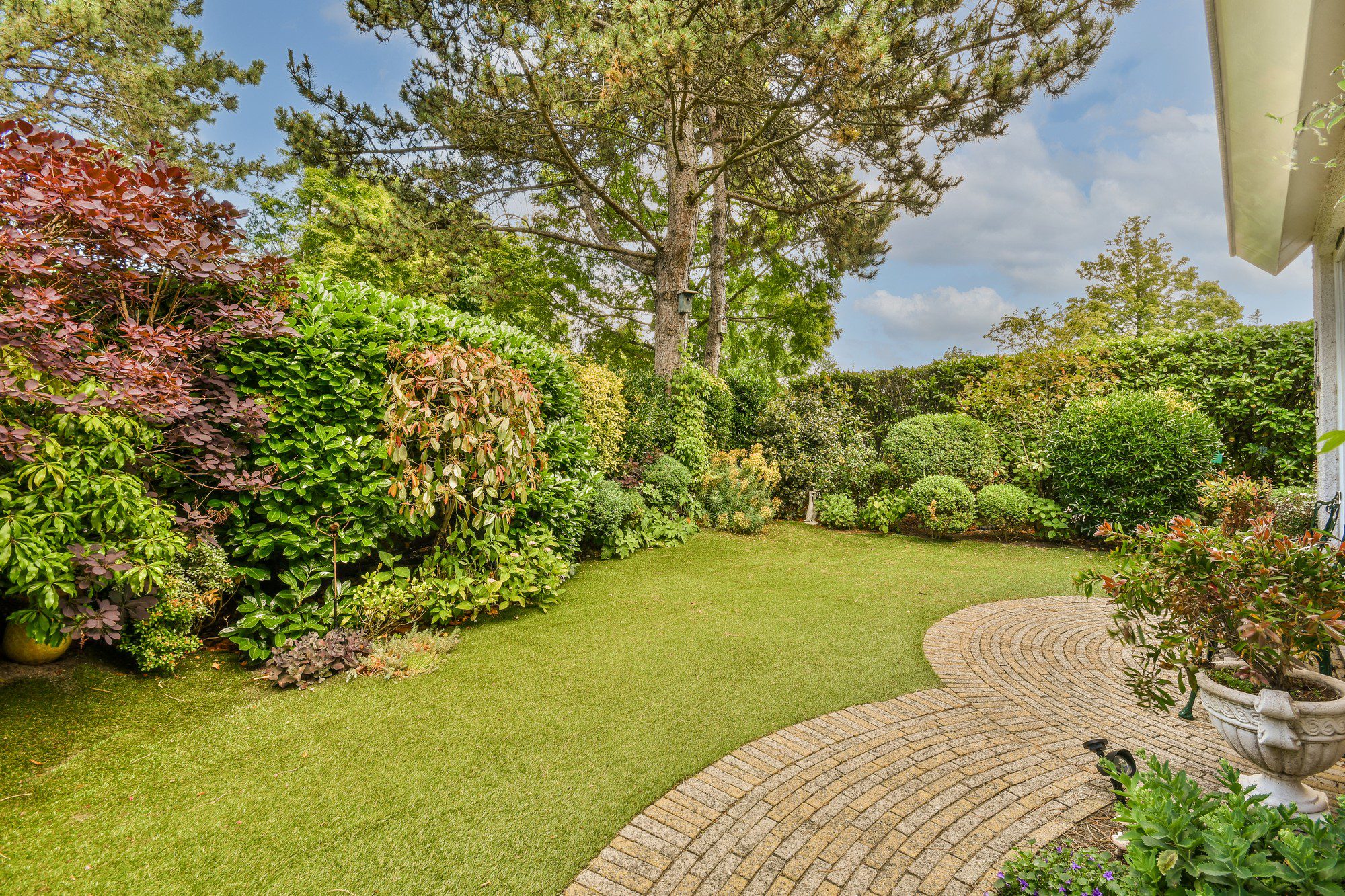 The image shows a well-manicured garden with a variety of plants. In the garden, there are shrubs and trees of different shapes, sizes, and colours, providing a diverse and lush landscape. The garden features a neatly trimmed lawn with a curved brick pathway leading through it, indicating careful maintenance. Surrounded by hedges and greenery, the garden presents a private and serene outdoor space. Additionally, there's a part of a building visible to the right side of the image with what appears to be a patio or covered area, which suggests this garden is part of a residential property. The sky is partly cloudy, indicating fair weather, and the lighting suggests it might be daytime. The overall impression is of an inviting and peaceful garden setting.