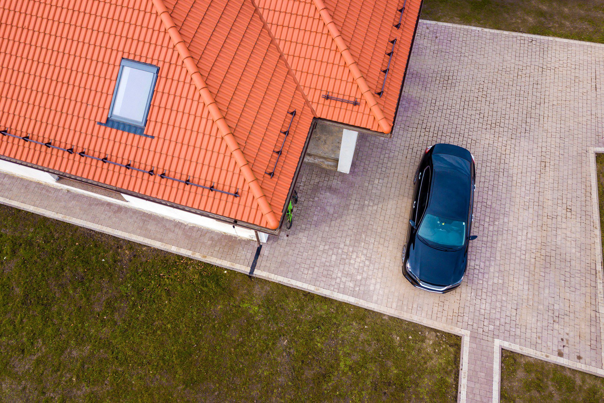 This image shows a top-down aerial view of a residential area. In the centre of the frame, there's a dark-coloured car parked on a brick-paved driveway next to a house. The roof of the house is covered with orange tiles and has a single window visible. To the left of the house, there's a grassy area with a light green garden hose coiled up near the wall. The layout appears very neat and orderly, with the driveway and adjacent pathways having a uniform brick pattern.