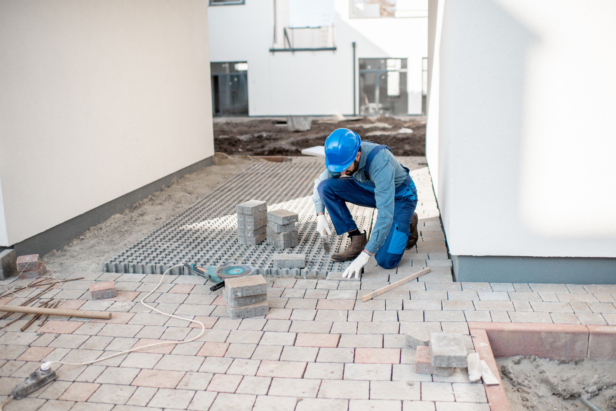 The image shows a construction scene where a worker wearing a blue hard hat and blue coveralls is installing paving stones or bricks to create a walkway or patio area. They are kneeling on the partially completed pavement, actively laying down the bricks. The ground preparation includes a layer of gravel, and there is a pattern to the brick placement, indicating a particular design for the walkway. Some tools and materials are scattered around the site, including what appears to be a level or straightedge, additional paving stones, and possibly a cutting tool for shaping bricks. The background suggests this is part of a larger construction area or building complex, as there is construction debris and unfinished landscaping visible. The worker is focused on their task, demonstrating manual labour typically involved in such construction projects.