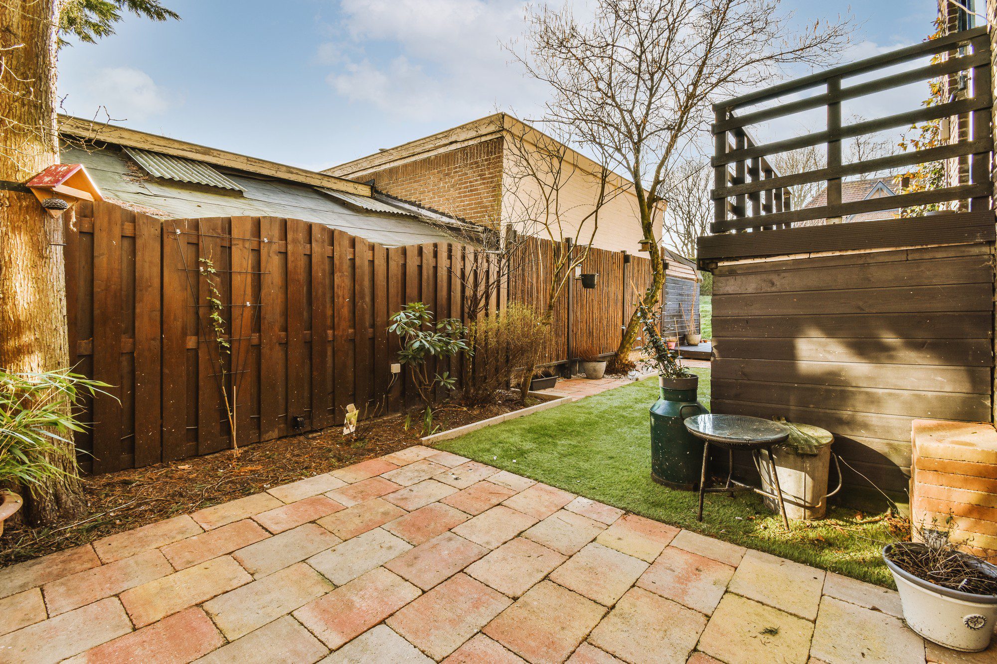 The image displays a well-kept backyard with various features. There is a terracotta-coloured paving stone patio area that leads to a section of artificial green grass. Surrounding this area are wooden fences on two sides providing privacy, and at the back, we can see a corrugated metal shed with its roof sloping downwards. There's an array of plants and shrubs planted along the edge of the fences, creating a garden border. On the right, there is an outdoor structure with wooden slats that might function as a privacy screen or a support for climbing plants. Accessories like a watering can and metal basin suggest the space is used for gardening activities. The setting appears to be in a temperate climate, given the look of the garden and the leafless tree hinting at a season outside of summer. The sun casts shadows indicating either morning or late afternoon light.