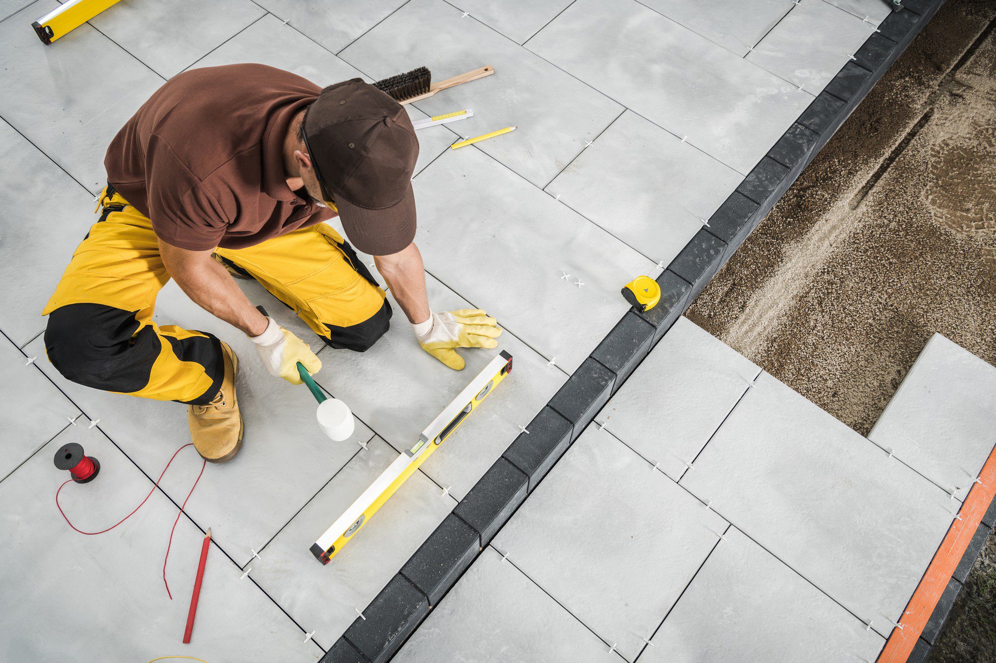 The image shows a person engaged in construction work, specifically laying tiles. The individual is wearing a brown cap, a brown shirt, yellow work pants with knee pads, and safety gloves. The person is kneeling on the gray concrete tiles that have been partially laid, with some areas still incomplete, showing the substrate or base layer where further tiles will be placed.The person appears to be applying or preparing to apply an adhesive using a notched trowel, which is a tool commonly used to spread adhesive in a uniform pattern before laying tiles. There are spacers placed between the tiles to keep them evenly spaced, ensuring a consistent gap for grout.In the surroundings, there are several tools visible, indicating the various stages of tile work: a measuring tape, a spirit level for checking flatness and alignment, a red wire or string that may serve as a guide for laying tiles straight, and a broom handle suggesting ongoing or completed cleanup. The area of work is bordered with a black flexible edge restraint, which might be part of the design or used to contain the substrate.Overall, the image captures a moment of manual labour in the construction or renovation industry, with a focus on tile installation.