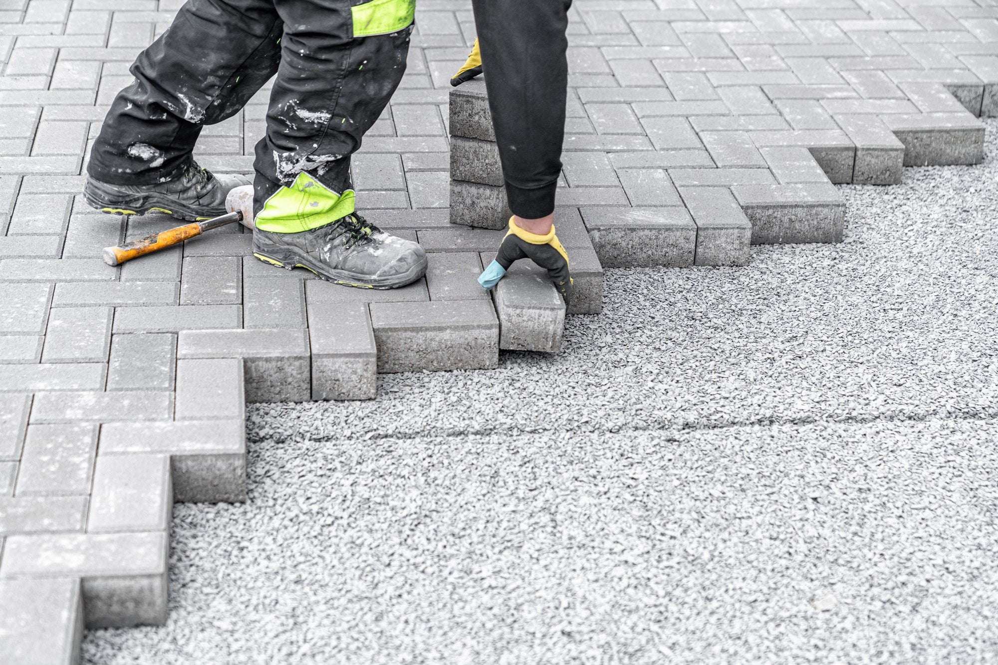 The image shows a person engaged in laying paving blocks or bricks. The individual is wearing work pants and high-visibility clothing, which suggests they are a construction worker or laborer. In front of them is a surface covered with a layer of fine gravel or aggregate, which serves as a base for the paving stones. They are holding one of the blocks with gloved hands, preparing to set it in place. To the left, there is a rubber mallet resting on the incomplete paved surface, likely used to tap the blocks into precise alignment and ensure a flat surface. The paving pattern looks to be a simple, staggered linear design.