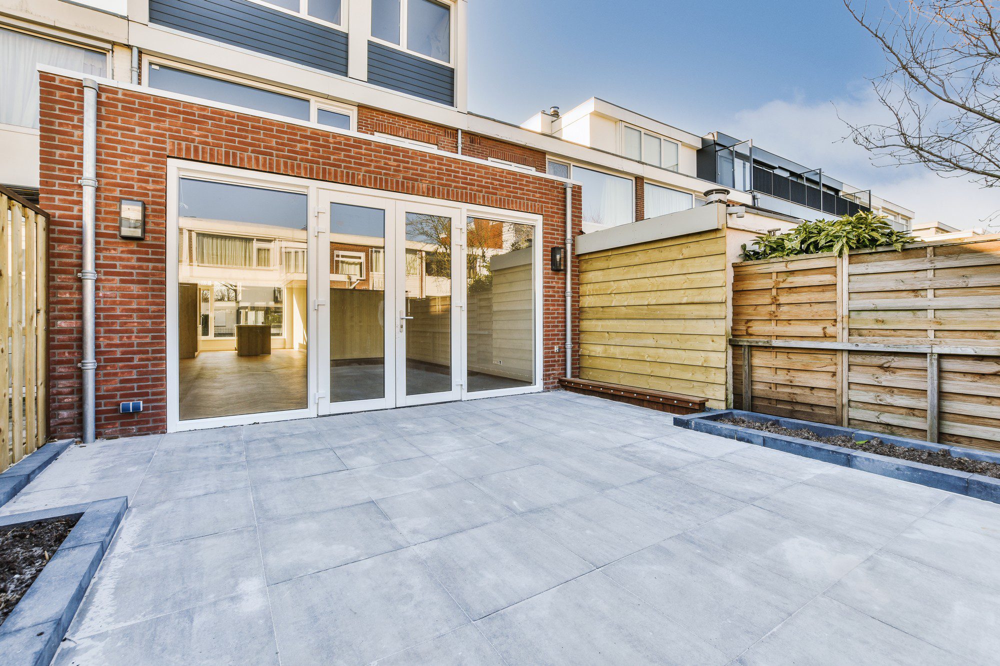 The image shows the outdoor patio area of a modern house during the daytime. The patio is constructed with large, square, smooth paving stones. To the right, there is a wooden slat fence, providing privacy to the area. The house itself has a combination of brickwork and siding, with large glass doors and windows that open onto the patio, offering a view into the interior living space of the home. Above the ground floor, you can see the upper story of the house with a balcony that features a glass balustrade. The sky is clear and blue, suggesting fair weather, which complements the outdoor living design of this space.