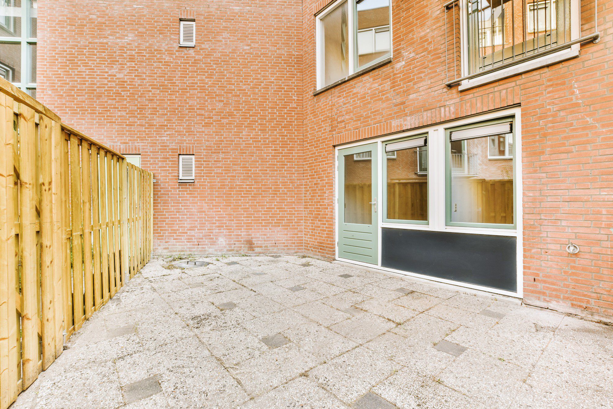 The image shows an outdoor area with a paved floor, which looks like a small courtyard or a back alley of a residential or commercial building. There is a red brick wall with several windows and a narrow, small window that seems to have vents, possibly for air exchange. On the left, there is a wooden fence that appears to be the boundary for the area.On the ground level of the brick wall, there's an entrance consisting of a light-coloured door flanked by two tall windows, which are fitted with reflective glass. Above this entrance, there is a barred window for security or safety. This setup suggests the image might be from the back of the building or a less public facing area, possibly designed for privacy and functionality rather than aesthetic appeal.The overall appearance suggests a modern, utilitarian design possibly found in urban or suburban areas. The space could serve as a small private outdoor area for residents or employees, or possibly as a service entrance.