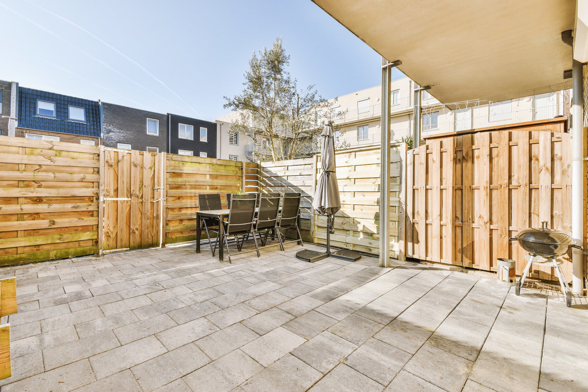 This image shows a residential backyard patio area. The patio floor is made up of large square paving stones. There is a wooden fence enclosing the space, providing privacy, and a wooden gate can be seen at the centre of the fence. Within the patio, there is a set of outdoor furniture that includes a table and four chairs, suggesting a space for dining or relaxation. An upright parasol is beside the table, currently closed, and to the right, there is a kettle-style charcoal grill. Off to the side, there's also a small tree and the edge of what appears to be either a shelter or a part of the house extending over the patio, potentially providing some shade. The weather seems to be sunny and the sky is clear. The buildings in the background have a modern architectural style.
