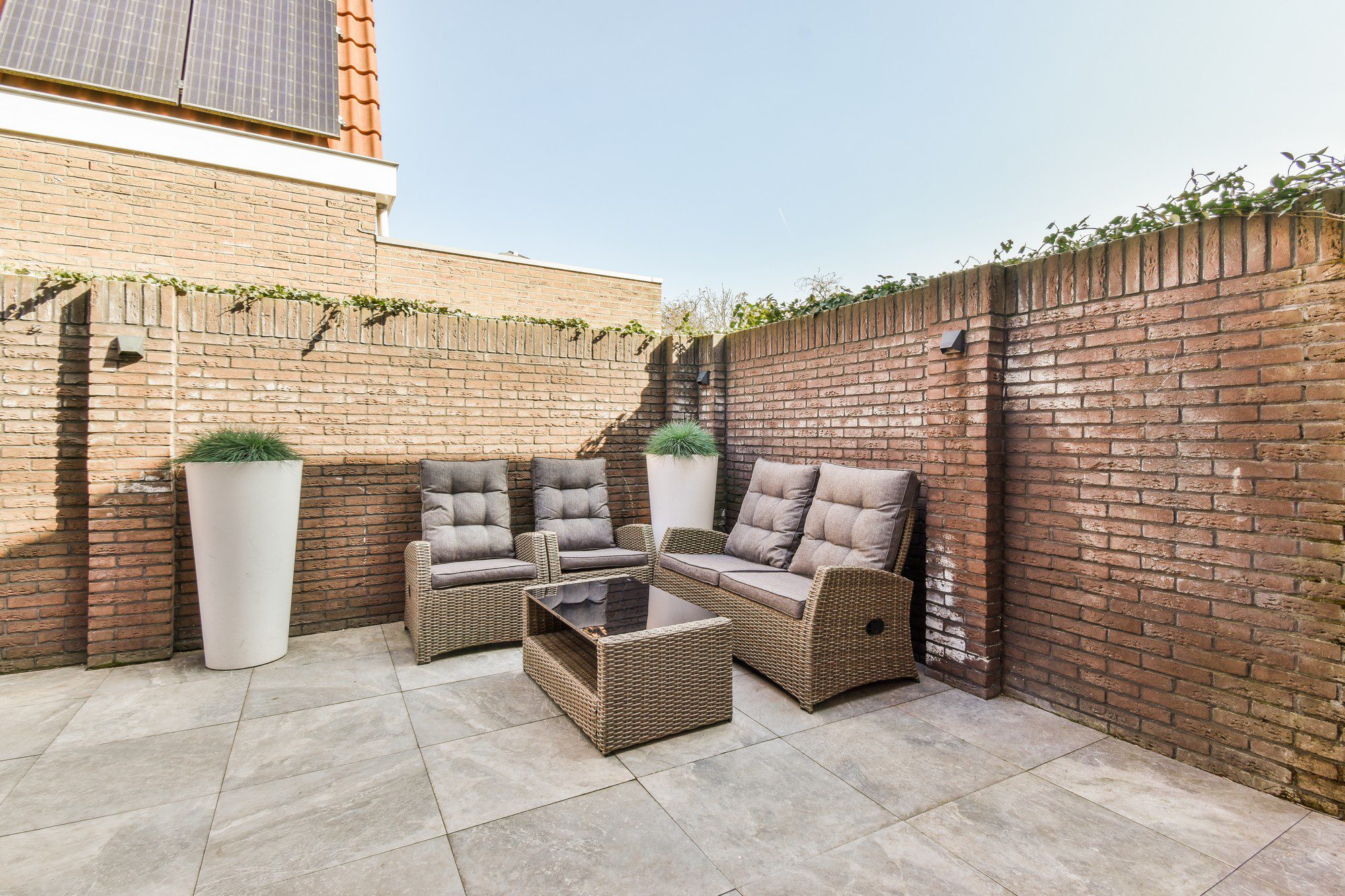 This image shows an outdoor patio area enclosed by brick walls. There is a set of modern patio furniture that includes two single armchairs, one double sofa, and a low table, all made with woven materials resembling rattan and topped with gray cushions. There's a tall white planter with a green plant in the corner. The floor is tiled with large, smooth paving stones, and there are climbing plants along the back wall. The sky is clear, indicating a sunny day, and the sun is casting shadows on the walls, highlighting the texture of the bricks. The corner of a building with a red roof and solar panels is visible at the top left corner, suggesting the patio might be adjacent to a residential home.