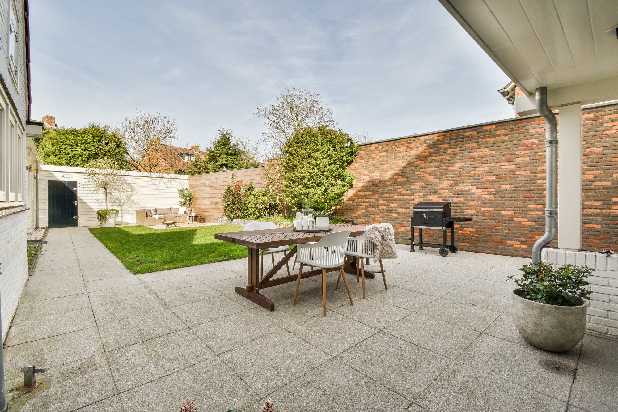 The image shows a well-kept outdoor patio area of a residential property. Key features include:- A spacious lawn with neatly cut grass.
- A patio dining set composed of a wooden table and chairs, suggesting a space for outdoor dining or relaxation.
- A barbecue grill positioned to the right, indicating a place for outdoor cooking.
- A large brick wall that encloses the area, providing privacy.
- A variety of shrubbery and a tree, which add greenery and life to the space.
- Part of a house with a downspout visible, showing that the patio is adjacent to the structure.
- A paved walkway leading up to the patio area, providing a clean and easy path to navigate through the garden.
- Potted plants adding decorative elements to the space.