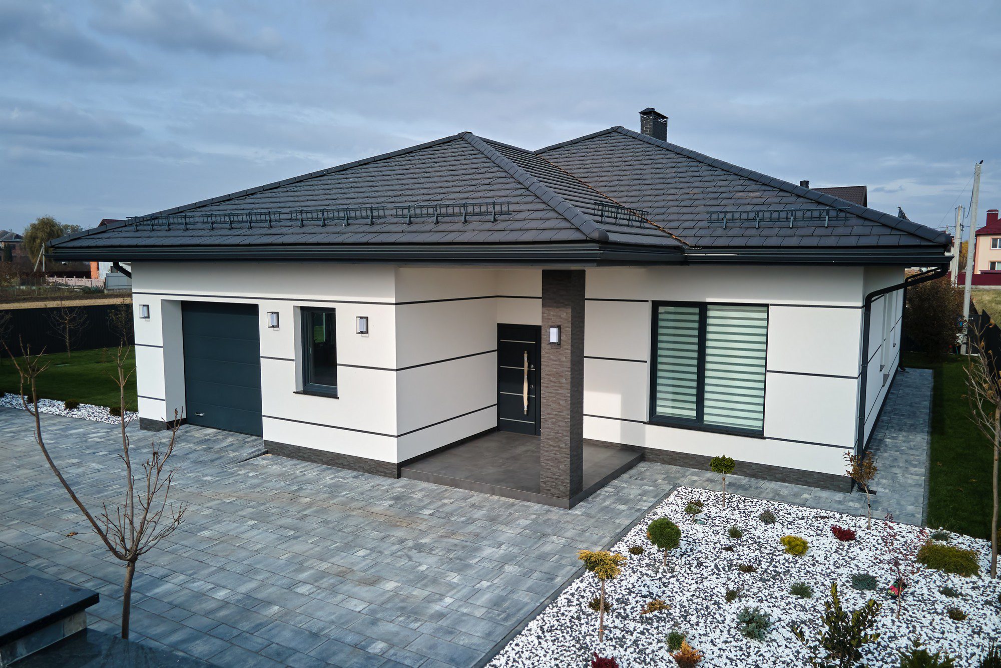 This is an image of a modern single-story detached house with a hip roof. The facade of the house features a combination of white render and dark gray or black cladding. The home has dark-framed windows, some of which are large and likely let in plenty of natural light. There is a dark entrance door complemented by a matching garage door.The house is surrounded by a well-maintained outdoor area with a paved driveway and walkway, and there is a landscaped garden featuring a mix of flowering plants, shrubs, and decorative stones. There are no people or animals in the image; the focus is on the architecture and landscaping of the property. It looks well designed and modern, with attention to curb appeal and outdoor aesthetics.