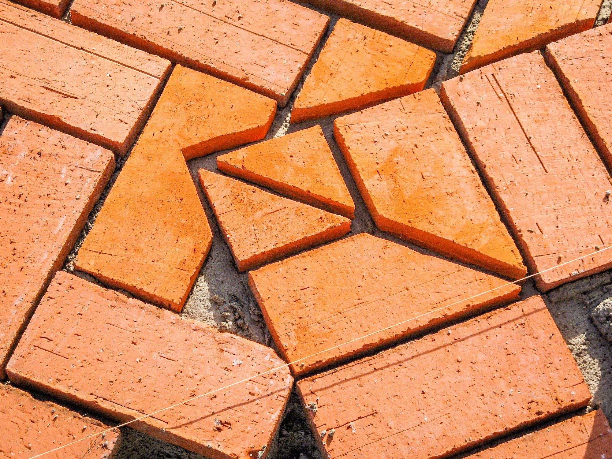This is an image showing a number of red bricks that are scattered and not laid in a structured or completed pattern. Some bricks appear to be partially embedded in a gray substrate, which might be concrete or mortar, and thin lines of what looks like string lines are visible on the surface, possibly used to align the bricks during the process of paving or constructing a pathway, patio, or brick wall. The image captures the texture and colour of the bricks, highlighting the sense of an unfinished or in-progress brick laying project.