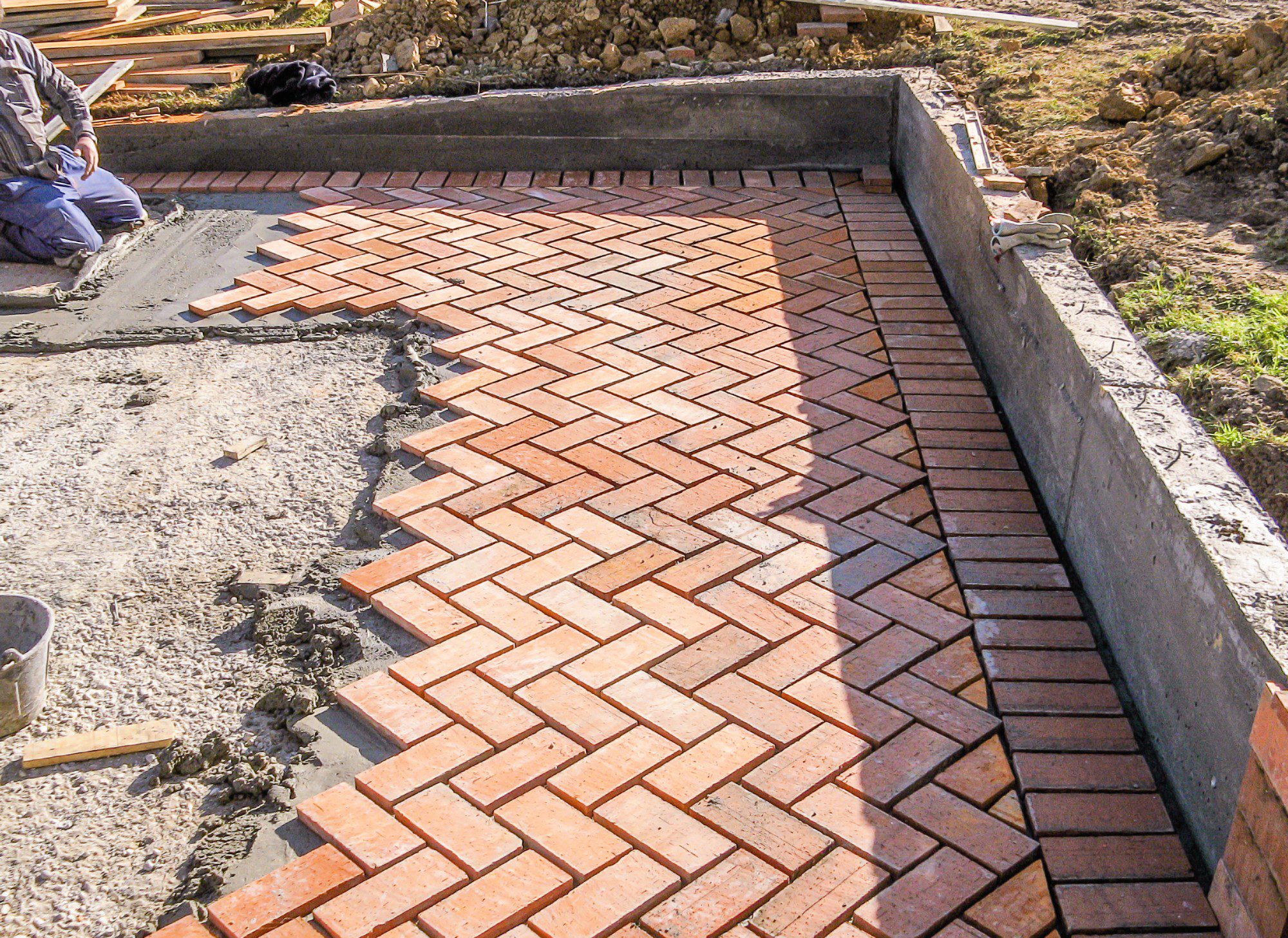 The image shows a construction or renovation site where brick pavers are being laid in a herringbone pattern to create a hard, durable surface, likely for a patio or a walkway. A concrete retaining edge surrounds the area where the bricks are being installed, which helps to keep them in place and maintain the structure's shape. There is construction material and debris around the site, including a bucket, piles of soil, and other building materials. In the background, there appears to be a person sitting on the ground wearing work clothes, possibly taking a break or assessing the work done so far, and next to them is a calm black dog lying on the ground. The unfinished area with exposed concrete suggests that the work is still in progress.