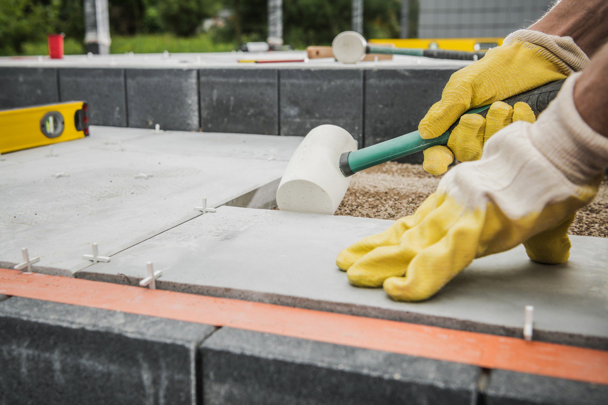 The image shows a construction or landscaping scene where a person wearing yellow work gloves is using a rubber mallet to adjust or set a paving stone into place. The individual is in the process of laying or installing pavers on a base of sand or fine gravel. You can also see spacer crosses between the slabs to maintain equal distances for the joints. We can partially see a spirit level on the left corner of the image, indicating that the person is ensuring that the pavers are level. The backdrop includes some greenery, suggesting an outdoor setting, and there's a construction block wall that possibly forms part of the structure or design they're working on.