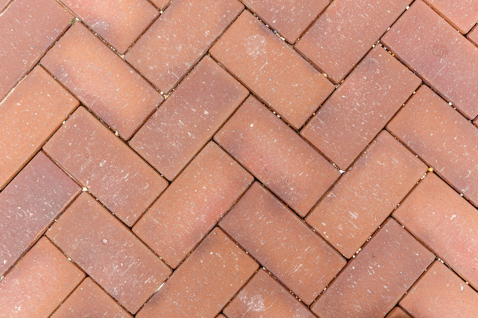 This image shows a surface covered with interlocking brick pavers arranged in a herringbone pattern. The bricks are reddish-brown in colour and there are visible small gaps filled with sand or another fine material that helps to keep the bricks in place. The pavers are slightly worn, indicating that they have been walked on or used for some time. The herringbone pattern is a popular design for outdoor patios, walkways, and driveways due to its pleasing aesthetic and stability under foot traffic.