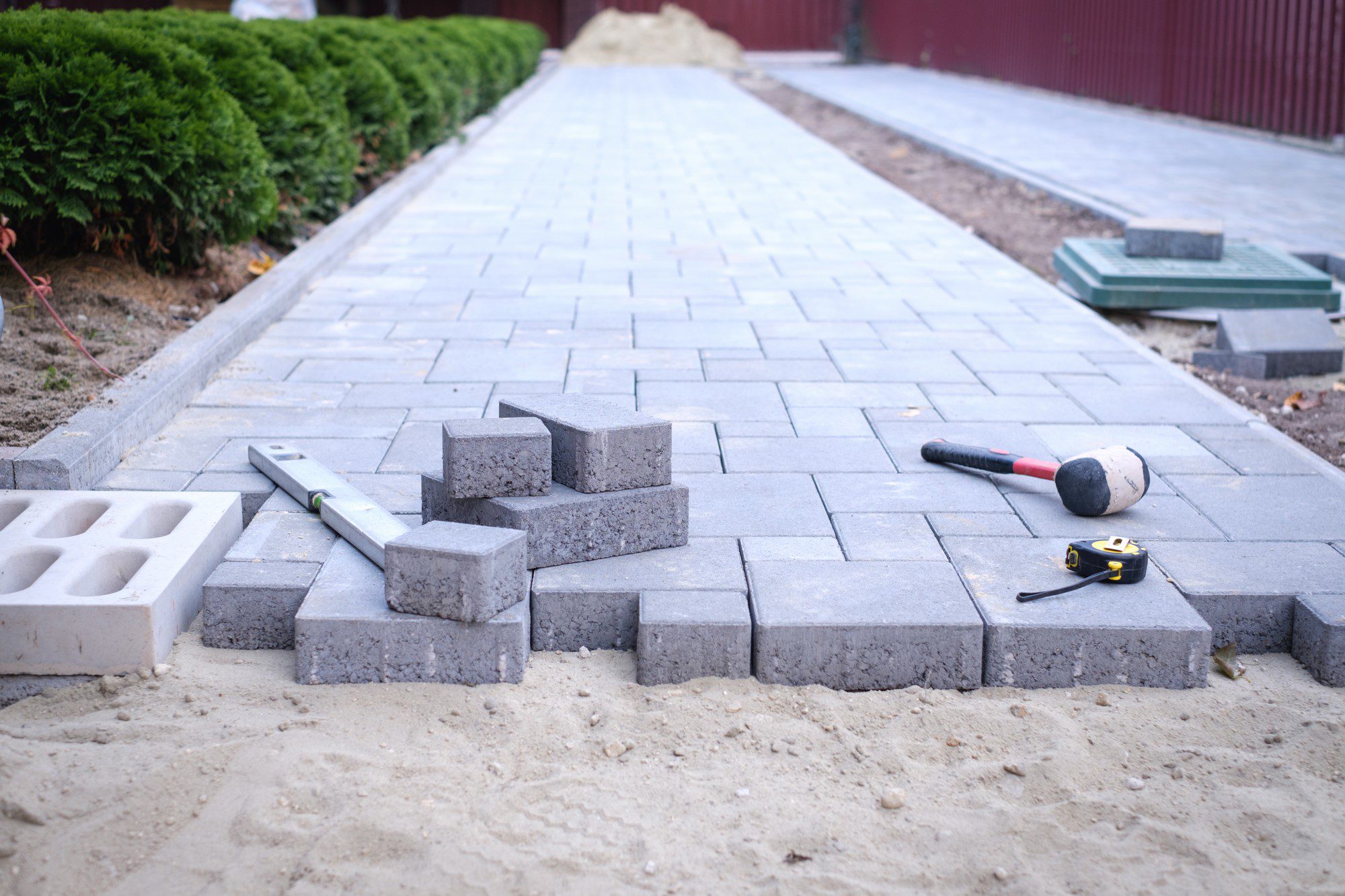 This image shows a work in progress of laying paving stones to create a pathway or patio. The individual gray paving stones are neatly arranged on the ground, forming a pattern on an area of compacted sand. To the side, some stones have been stacked up, likely in preparation to be placed. The scene includes a few tools commonly used in this type of work: a rubber mallet, presumably used to gently tap the pavers into place; a spirit level for making sure the pavers are even and level; and a tape measure for precise measurements. In the background, you can see a lined garden bed with shrubs and a pile of sand, indicating that the construction work is ongoing. A metal or plastic border appears to edge the pathway, providing a clean line and helping to hold the pavers in place. The environment suggests residential landscaping or possibly public walkway renovation.