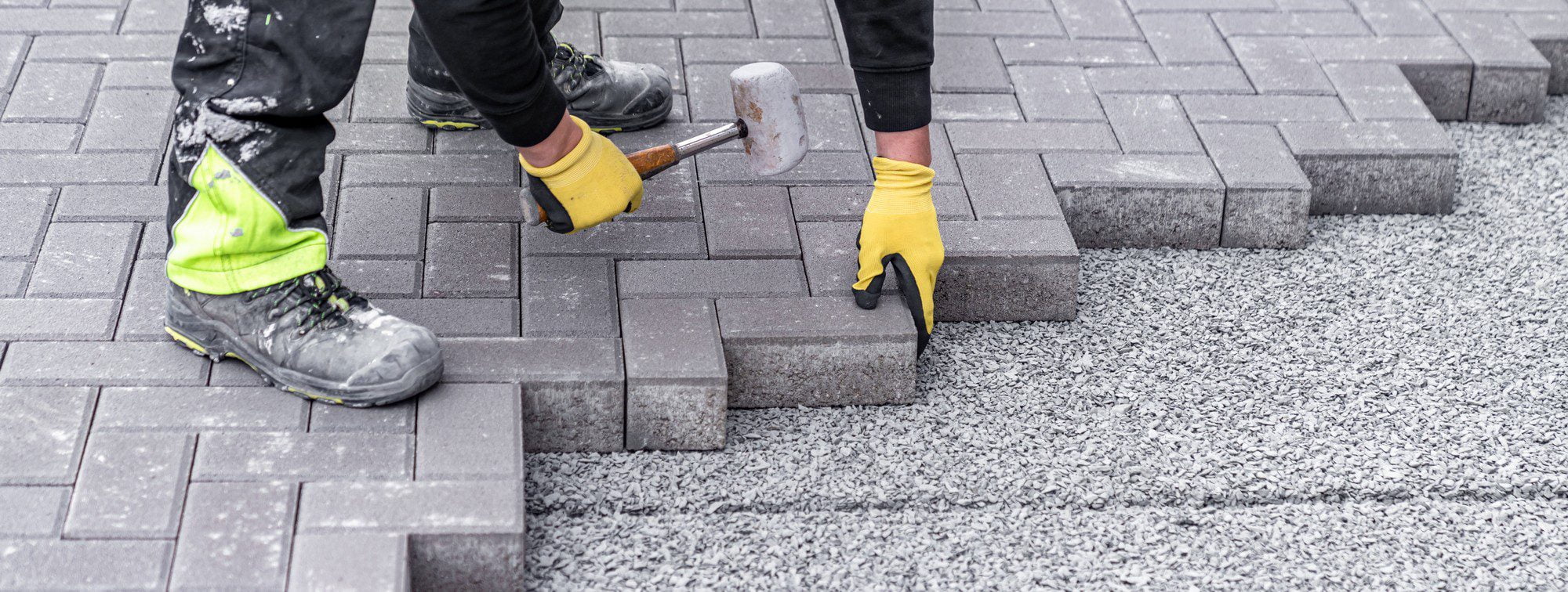 The image shows a person working on laying interlocking paving stones, commonly seen in driveway, sidewalk, or patio constructions. The worker is wearing safety gear, including a pair of gloves and a high-visibility stripe on their boot, which suggests adherence to safety regulations typically seen at construction sites. In their hands, they are holding a rubber mallet and paving stone, indicating they are in the process of installing or adjusting the pavers into position. The area that has yet to be paved is filled with a layer of gravel, which is used as a base material for the pavers to ensure proper drainage and a stable surface. The worker’s stance and the arrangement of the materials suggest they are focused on precision and quality in their work.