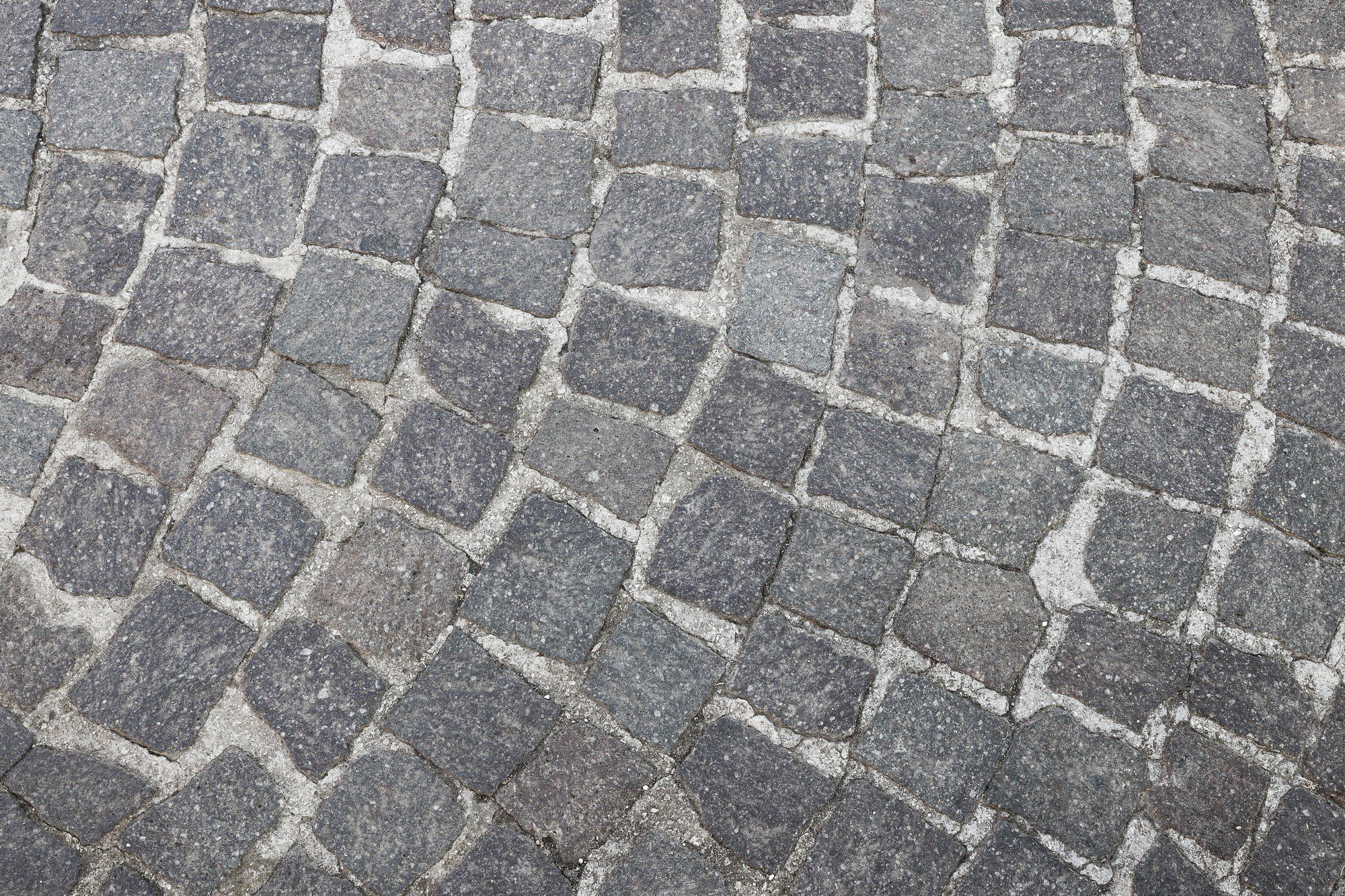 The image shows a close-up view of a cobblestone pavement. The stones are arranged in a somewhat staggered pattern, with lines of smaller, rectangular stones creating a border or pattern within the larger cobblestone layout. The colours of the stones are mostly shades of gray, and the surface appears to be worn, suggesting that the pavement has been used for some time. Such cobblestone surfaces are common in historic or picturesque areas and are known for their durability and aesthetic appeal.