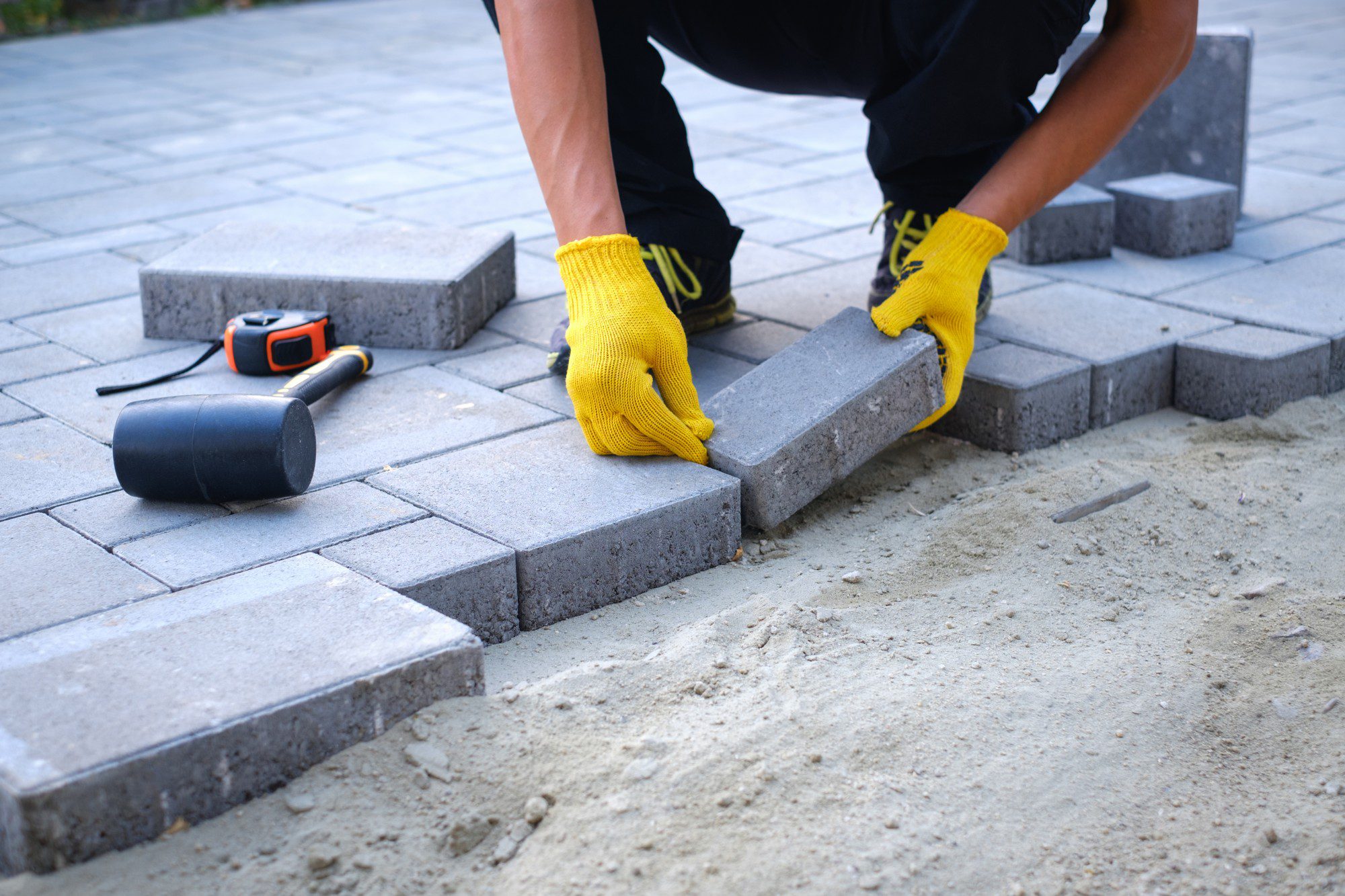 The image shows a person laying paving stones to create a path or pavement. The person is wearing yellow work gloves and is in the process of placing a gray rectangular paving stone onto the sandy ground. There is a rubber mallet and a tape measure lying on the already laid paving stones, indicating that the work is precise and requires measurement. The arranged stones create a pattern, and the unfinished edge suggests that the work is in progress. It's a snapshot of construction or landscaping work.