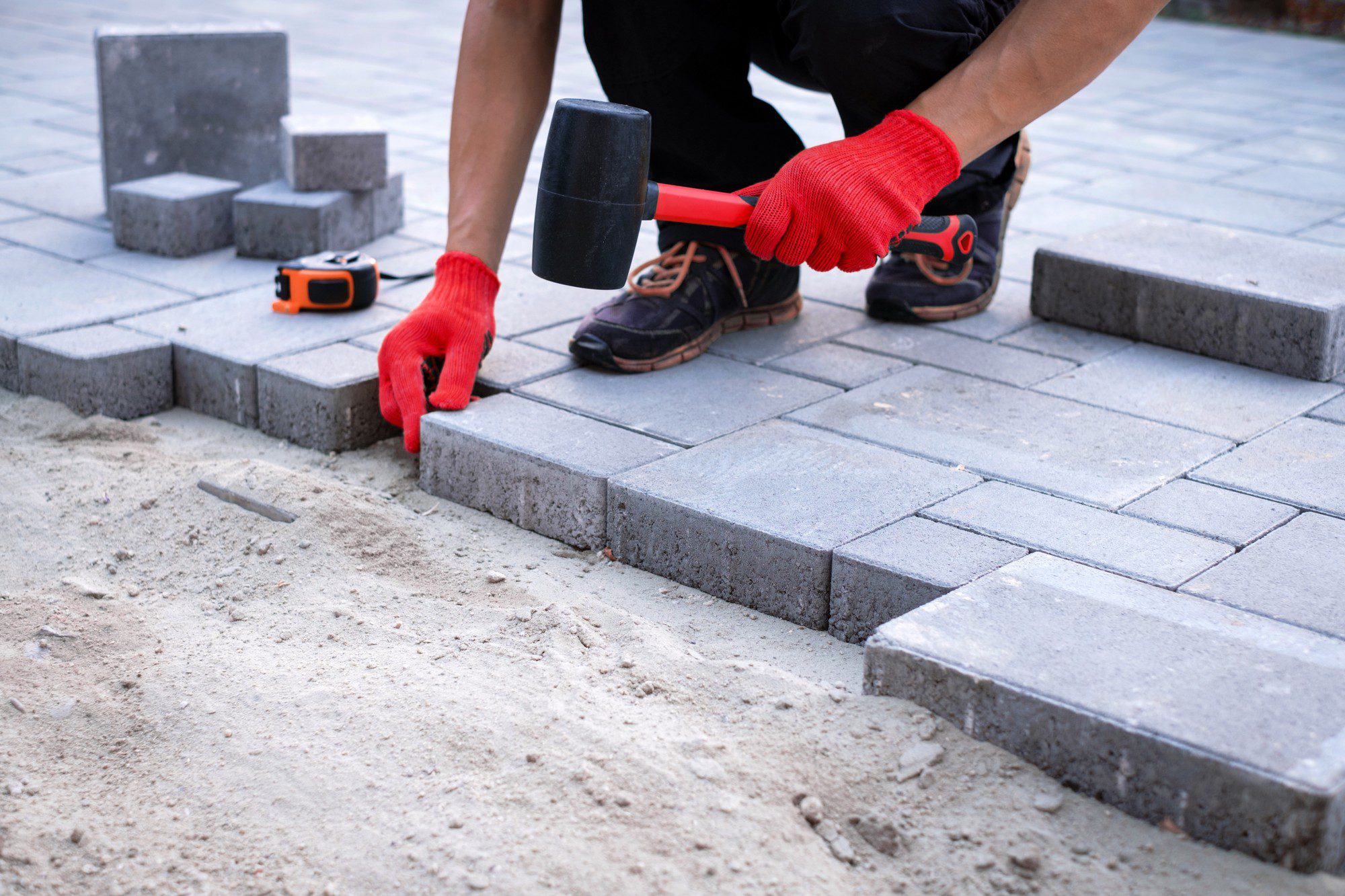 The image shows a person in the process of laying paving stones for a patio or walkway. The individual is positioned close to the ground, wearing work pants and shoes, and has protective red gloves on. They are using a rubber mallet, a common tool for this type of work, to ensure the paving stones are set at the correct level and are securely in place. To the left of the person, there is a tape measure, suggesting precise measurements are being taken during the installation process. The stones are gray and are laid out in an interlocking pattern, a method often used to create a stable and attractive surface. It appears to be an outdoor setting, possibly a construction or landscaping site.