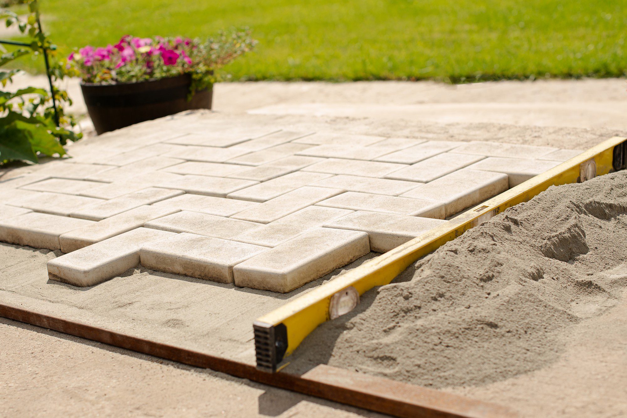 The image shows a paved area that appears to be in the process of construction. There are rectangular paving stones or bricks laid out in a herringbone pattern on a bed of sand. One of the paving stones is slightly raised above the rest, likely awaiting proper placement. A spirit level, which is a tool used to cheque surfaces for levelness, lies on top of the pavers, indicating that the work is being checked for precision. Some sand is piled to the side, indicating ongoing work, while the boundary of the paved area is defined by metal or wooden edging. The background shows a lush green lawn and a dark-coloured flowerpot with pink flowers, suggesting a residential garden setting. The sun is shining, and the overall atmosphere looks like a pleasant day for outdoor work.