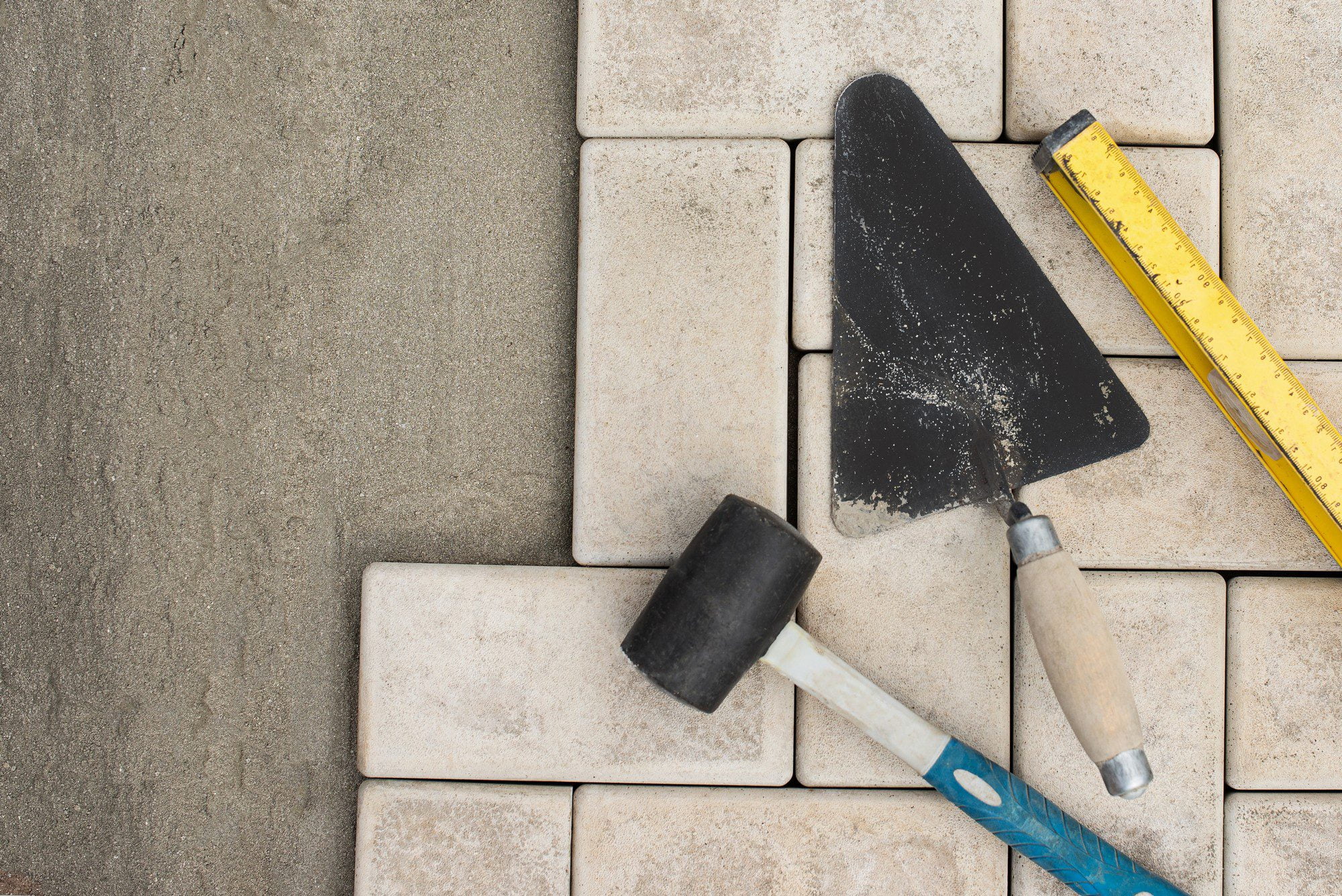 The image shows a collection of tools and materials typically used in laying or repairing paving stones. There is a rubber mallet, a trowel with remnants of a powdery substance (possibly sand or cement), and a yellow folding ruler. These tools are placed on top of and next to paving stones, some of which are laid down, while one seems to be partially lifted or not yet fully set in place. This suggests some sort of construction or maintenance work involving the paving stones is taking place.
