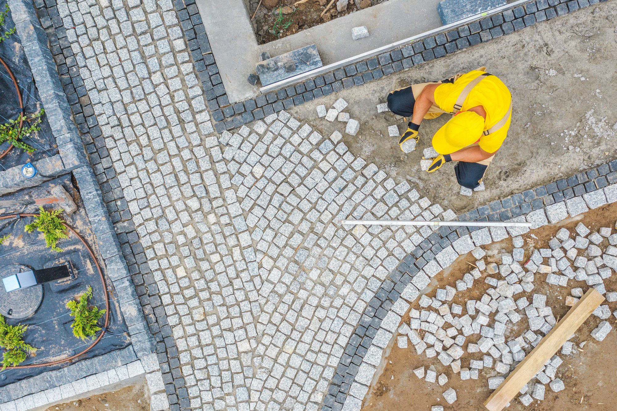 The image shows an aerial view of a person laying paving stones on a road or pathway. The person is wearing a yellow high-visibility jacket and a yellow hard hat, indicating that they are likely a construction worker engaged in manual labour. The pattern of the paving stones creates a herringbone design, and the worker seems to be meticulously placing the stones to maintain the pattern.You can see different shades of paving stones, with lighter ones forming the herringbone pattern and darker ones defining the borders and edges. There are unfinished areas with a base layer of sand or gravel visible, and loose paving stones are scattered around the work area, ready to be laid down. Also noticeable are elements of urban infrastructure, like a manhole cover which has been worked around, and conduit pipes visible in the excavated sections. The presence of a wooden plank suggests it may be used to help level or set the stones.It's a clear example of urban roadwork that involves significant attention to detail and craftsmanship.
