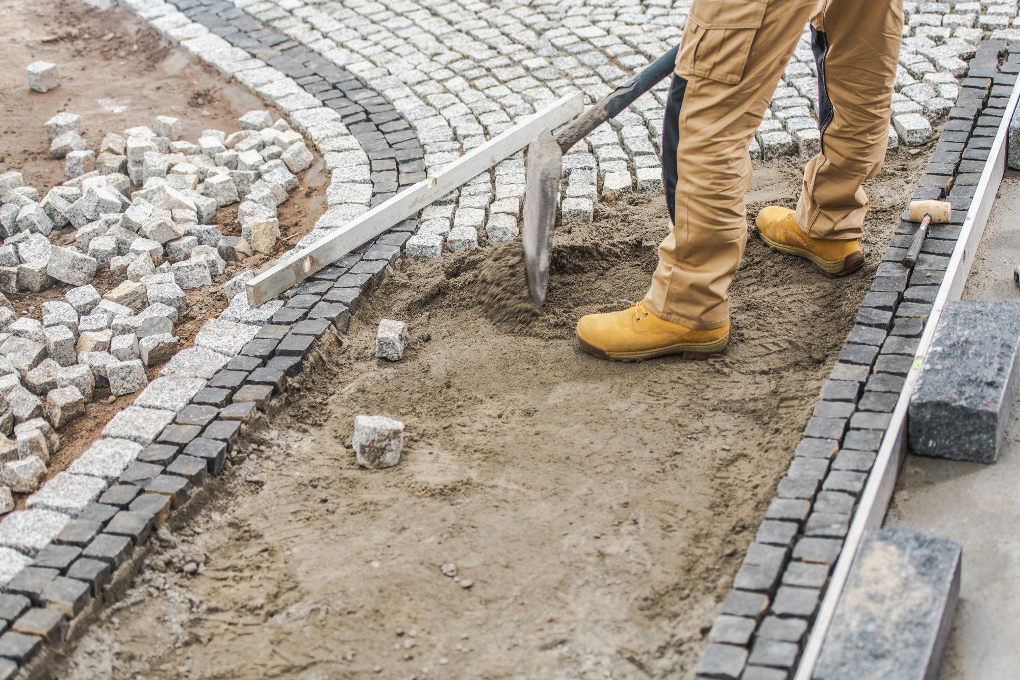 The image shows a person at work during the construction or repair of a cobblestone pavement. The person is wearing work boots and khaki pants, and they are using a shovel to spread what appears to be a layer of sand or fine gravel, which serves as the bedding for the cobblestones. You can see the unfinished section of the pavement where the base layer is exposed, along with a metal straightedge level resting on the surface to ensure the stones are laid at the correct height and alignment. Completed sections of the cobblestone path feature stones that have been neatly placed in an interlocking pattern. Some loose cobblestones are scattered around the work area, and the edge of the path is lined with a row of darker, larger stones or curb blocks, providing a clear border.