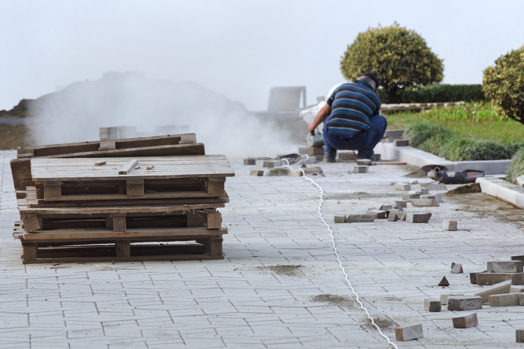 The image shows an outdoor setting with construction or maintenance work taking place. In the foreground, there are several stacked wooden pallets on top of a paved area. The paving stones give the impression that this could be a pedestrian path or patio.In the background, there is a person engaged in some kind of manual labour. This person is crouched over and appears to be handling or working with the paving stones, as suggested by the scattered array of stones and the dust surrounding the area of work. There is construction equipment on the ground next to them, which could be tools or machinery used for cutting or placing the stones.Near the person, white extension cords are visible, trailing across the ground and presumably connected to the equipment being used. The atmosphere seems to be dusty, likely from the work being done on the paving stones.Overall, the scene captures a moment of ongoing construction or improvement work to a paved outdoor area.