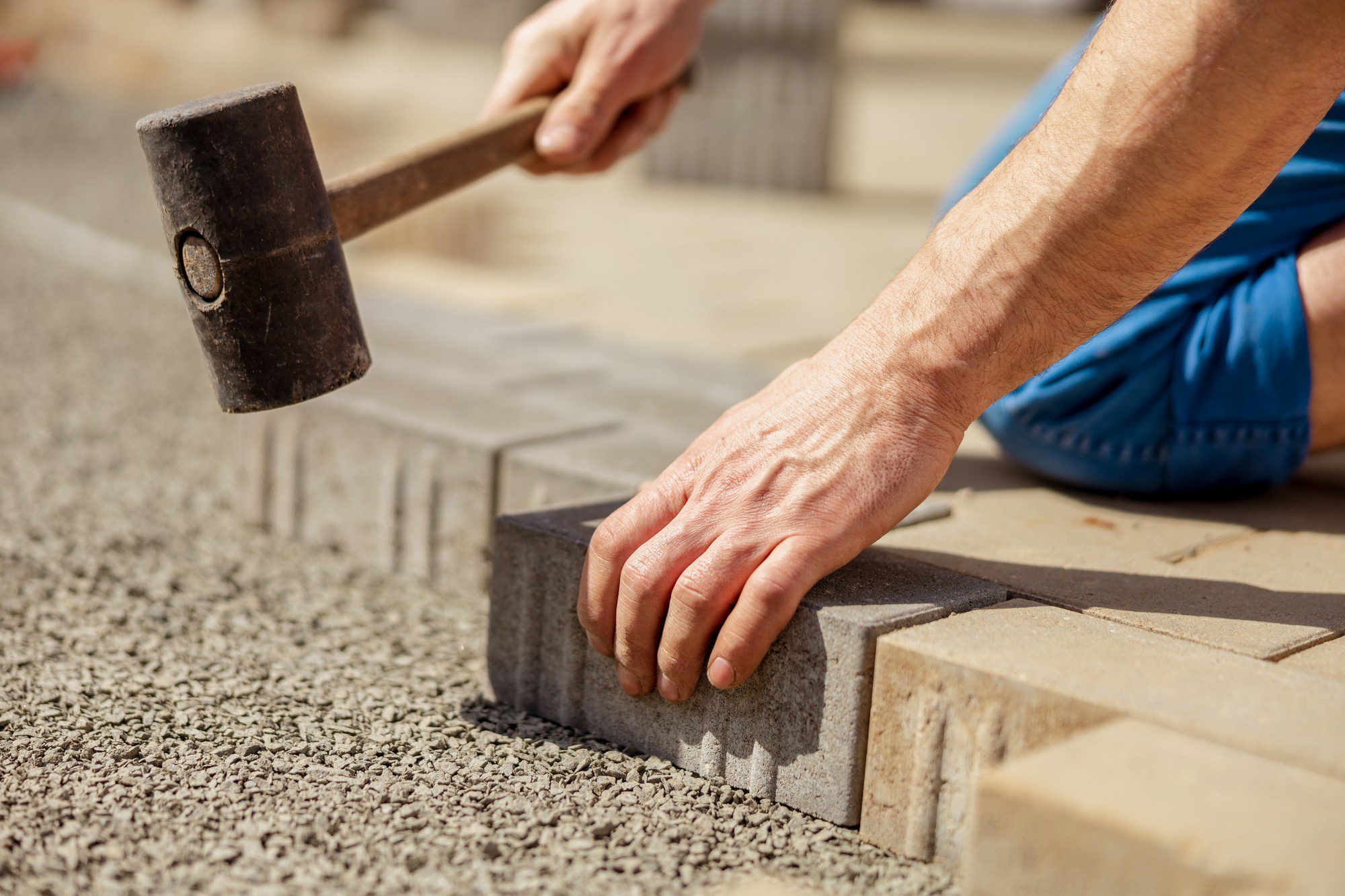 The image shows a close-up of a person laying paving stones. The person is using a rubber mallet to tap a paving stone into place on a bed of what appears to be a granular base, like sand or fine gravel. The person is wearing a short sleeve shirt and shorts, indicative of warm weather or outdoor working conditions. The focus is on the person's hands and the mallet, with the foreground and background slightly blurred to emphasize the action. This suggests the person is engaging in some kind of construction or landscaping work.