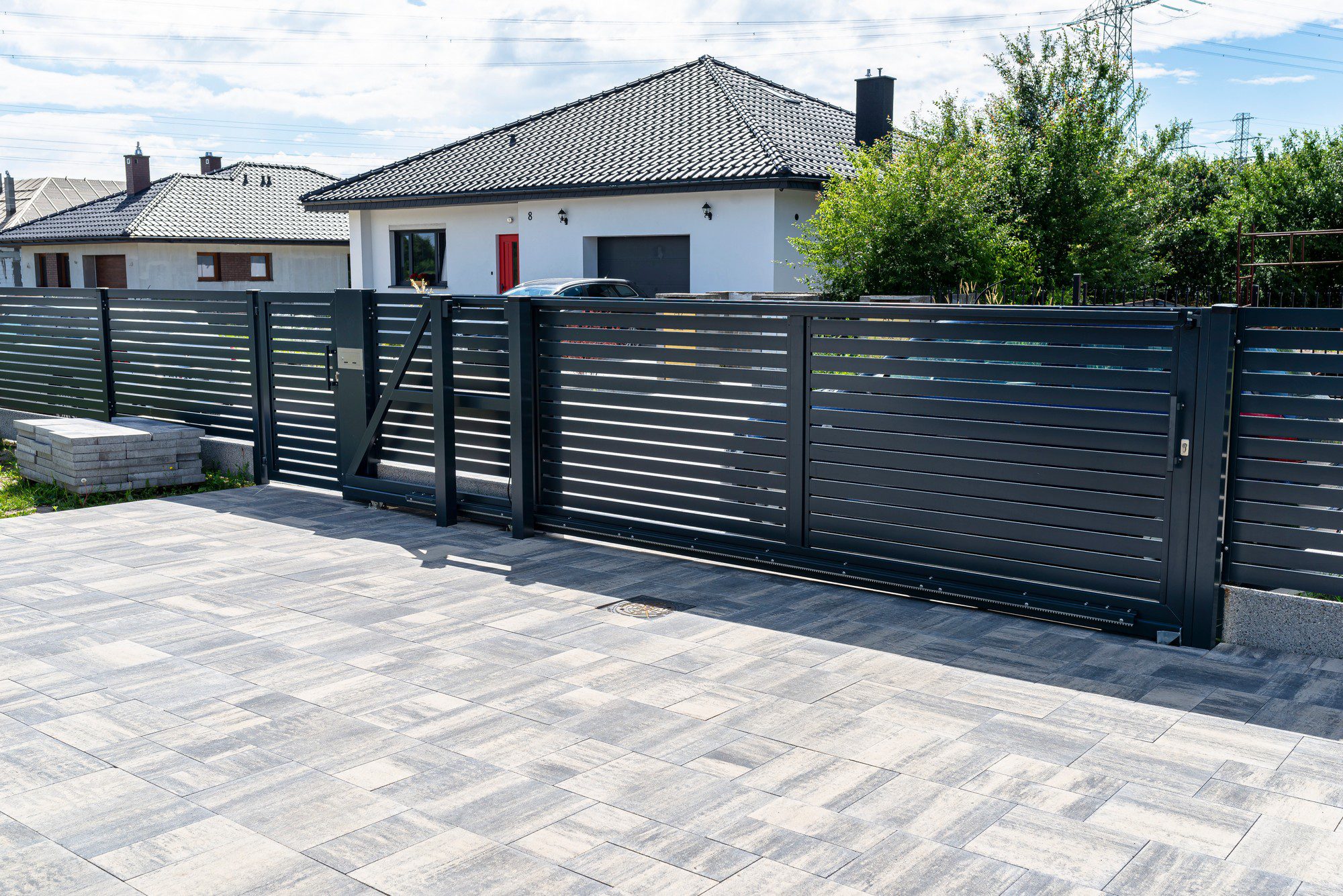 This image shows a modern residential setting featuring a stylish black metal fence with horizontal slats, providing privacy while also allowing some light and air through. There is a gate integrated into the fence, and it appears to be a sliding type given the track on the ground. The pavement in the foreground is made of rectangular paving stones laid in a repeating pattern. In the background, we can see the facade of a single-story house with a red door and dark tiled roof, which complements the contemporary aesthetic of the fence. There are also some green bushes and a clear blue sky, suggesting a pleasant, suburban environment.