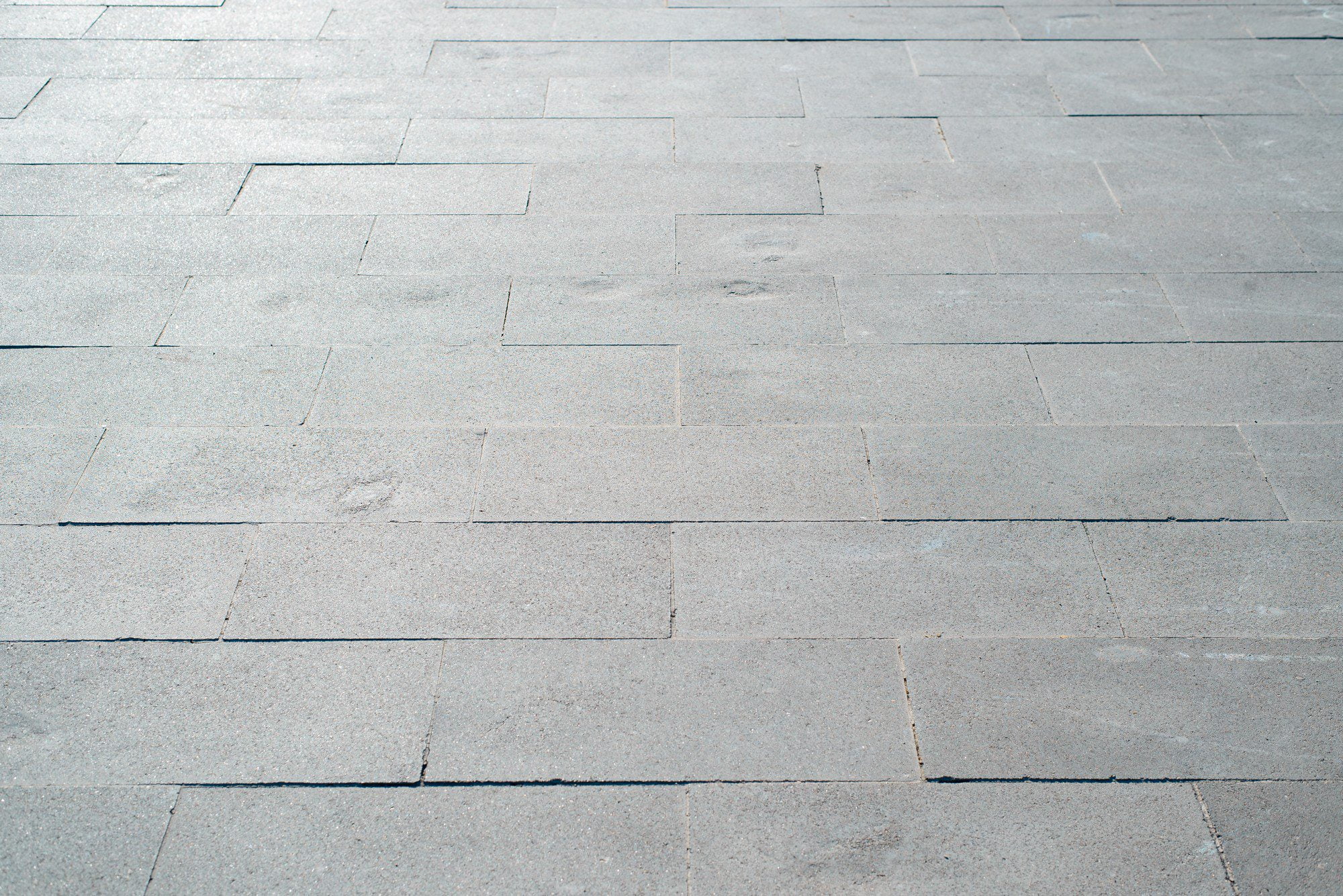 The image shows a paved surface consisting of uniform rectangular stone or concrete slabs arranged in a staggered pattern. The surface is flat and seems suitable for pedestrian traffic, indicating that it could be a section of a sidewalk, plaza, or other pedestrian area. The photo captures the texture and colour variations of the paving material under natural light, with shadows indicating a bright day. There are no discernible distinctive features or landmarks to indicate the specific location.