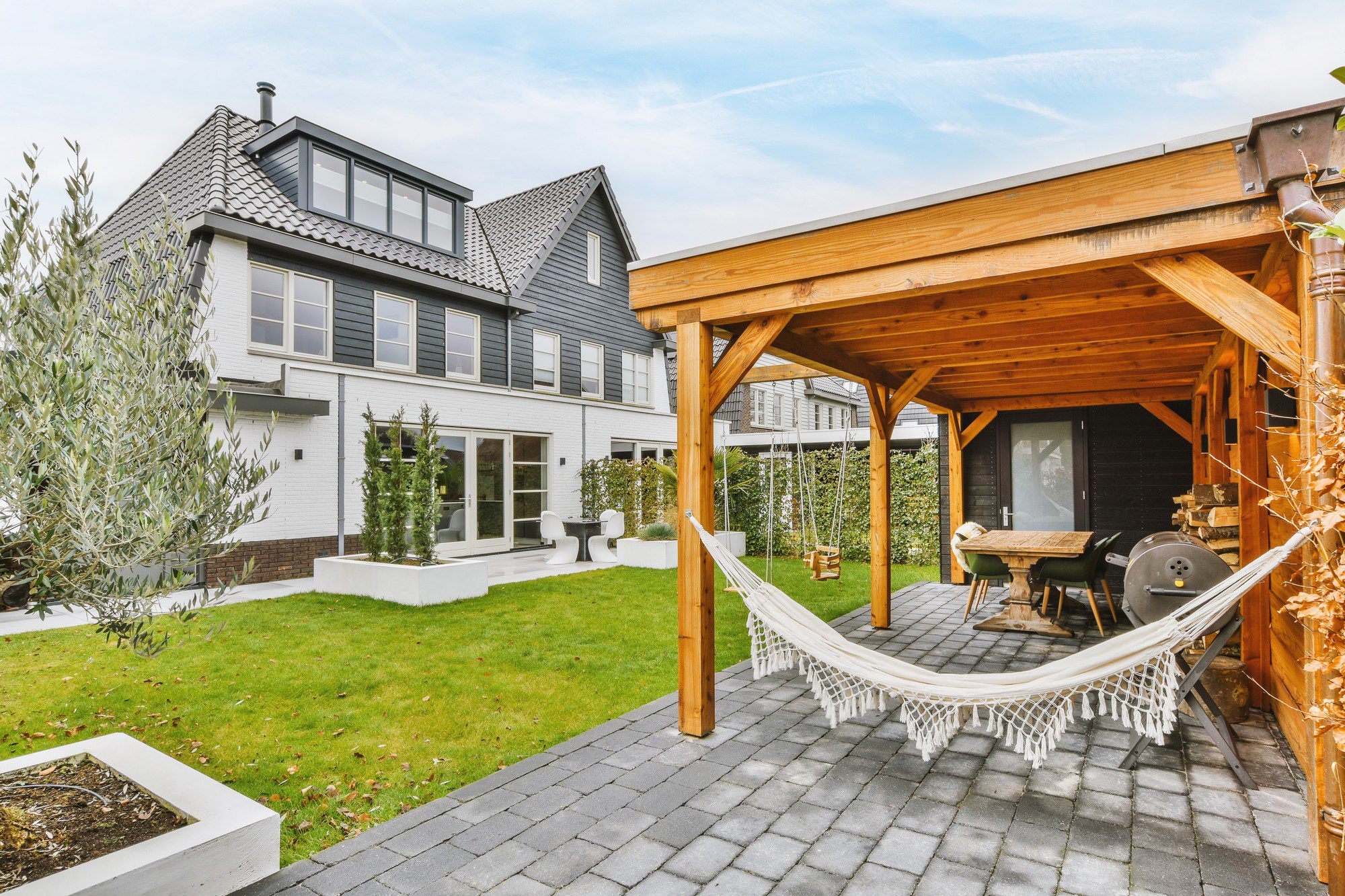 The image shows a modern two-story house with a mix of white and dark siding. There's an attached covered area with a wooden structure that provides a cosy outdoor dining and lounging space. A hammock is hanging in the foreground, adding a relaxed vibe to the setting. The backyard also features neatly trimmed grass, a well-maintained garden with a variety of plants in planters, and a paver stone patio. The house looks well-kept and the design indicates a blend of contemporary and traditional elements.