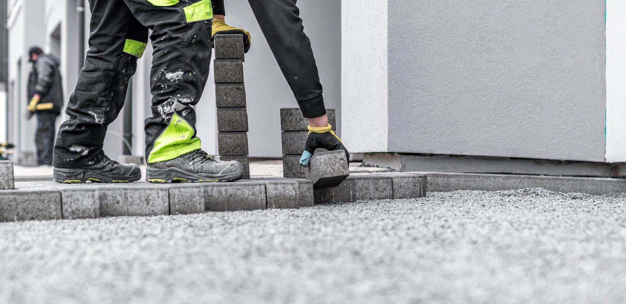 The image shows a close-up view of a construction or landscaping site where paving work is being carried out. There are two workers in the image. The one in the foreground is wearing dirty work trousers with reflective hi-visibility strips around the lower leg, and sturdy work boots, indicating that they are involved in manual labour. This person is stepping onto a raised platform or curb. The second worker in the background is bent over and appears to be handling materials or tools required for the job.Both workers are wearing gloves to protect their hands. The environment suggests an urban setting, perhaps a sidewalk or pedestrian area that is being paved or repaired. The paving blocks or stones being used are dark-coloured and stacked neatly in the foreground, with one being placed or adjusted by the worker. The ground is covered in a layer of gravel or small stones, which is typical for providing a stable base for paving. The building walls in the background indicate that this work is occurring near existing structures.