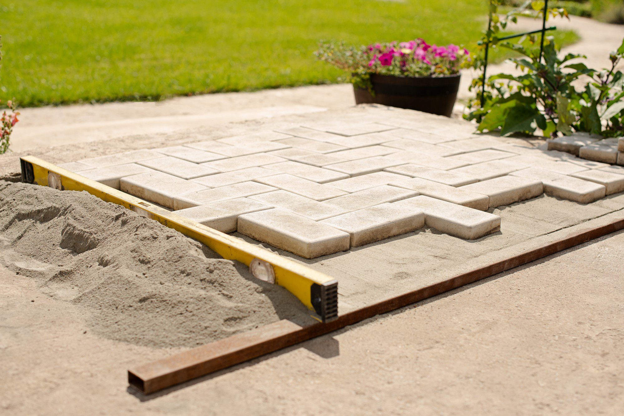 The image shows a section of a paved area, which appears to be under construction or renovation. There are uniform, rectangular paving stones laid out in a pattern on a bed of sand. Part of the pavement seems completed, while other stones are stacked and yet to be placed. A yellow level tool rests atop the stones to ensure that the surface is flat and even. On the left, there's a pile of sand, likely used as a base layer for the pavers. In the background, you can see a well-manicured lawn and a potted plant with pink flowers, indicating a garden or a landscaped area. The presence of the level tool and the orderly laying of the stones suggest that this is a carefully planned and executed landscaping project.