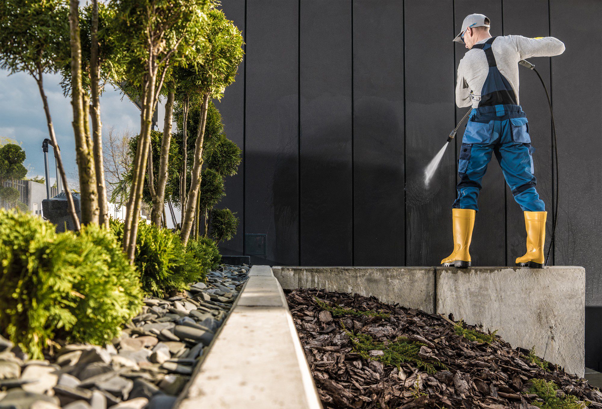 The image shows a person cleaning a black vertical surface using a high-pressure water spray. The individual is wearing a cap, a white long-sleeve top, blue work overalls, and yellow rubber boots. They appear to be standing on a concrete ledge or platform. To the left, there is landscaping with a variety of plants and shrubs, with a decorative layer of rocks and organic mulch. It looks like a maintenance or cleaning task being performed in an outdoor setting, likely as part of property upkeep. The high-pressure washer is creating a visible spray of water that is striking the wall and producing a streak of cleanliness against the darker surface, indicating a possibly significant contrast between the cleaned and yet-to-be-cleaned areas.
