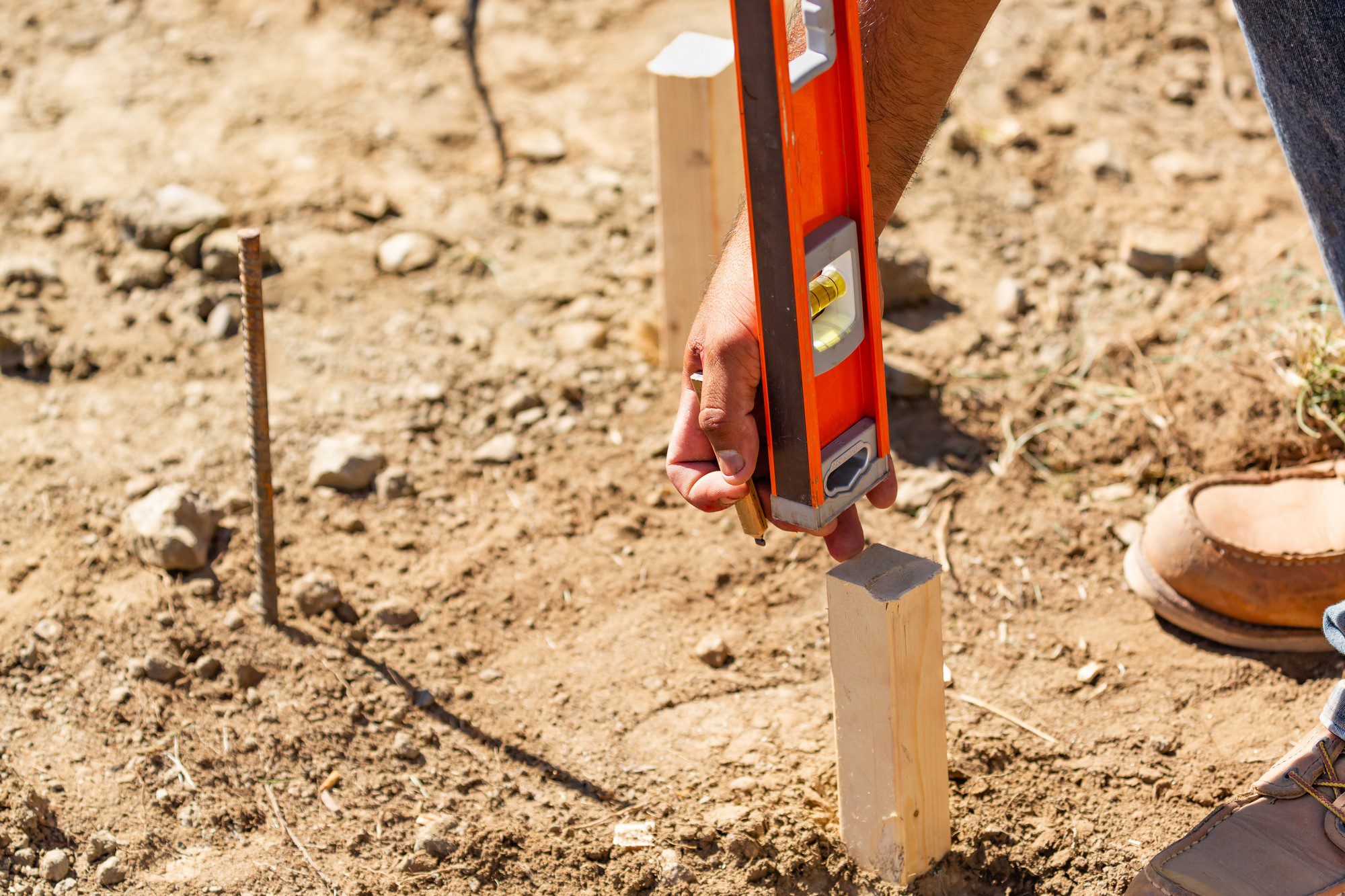 This image features a person using a red spirit level to cheque the verticality of a wooden stake that has been placed in the ground. Part of the individual's body is visible, specifically their hands holding the level and their lower legs; they are wearing jeans and brown boots. On the ground next to them is a rusty rebar or stake and a brown object that appears to be a bowl or hard hat. The ground itself looks dry and uneven with some rocks and sparse vegetation, suggesting an outdoor construction site or a similar work setting.