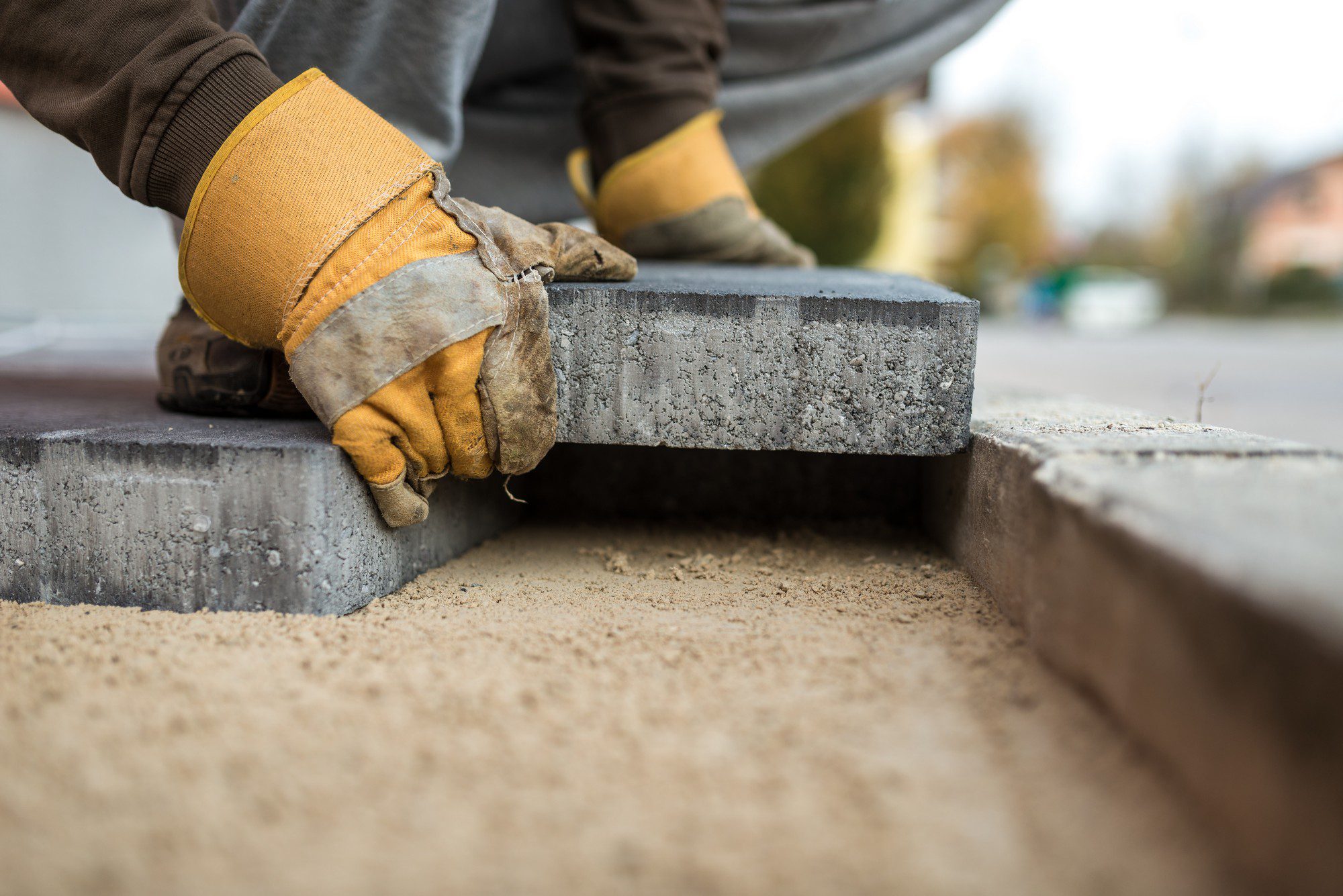 The image shows a close-up of a person's hands wearing heavy-duty, yellow work gloves while handling a concrete block or paver. The person seems to be working on paving or landscaping, as the concrete block is being placed on a bed of sand, which is typically done to ensure a level and stable base for pavement or hardscaping projects. The focus is primarily on the hands and the concrete block, emphasizing the manual and physical aspect of the work being done. The background is blurred, which helps to draw attention to the action taking place in the foreground.