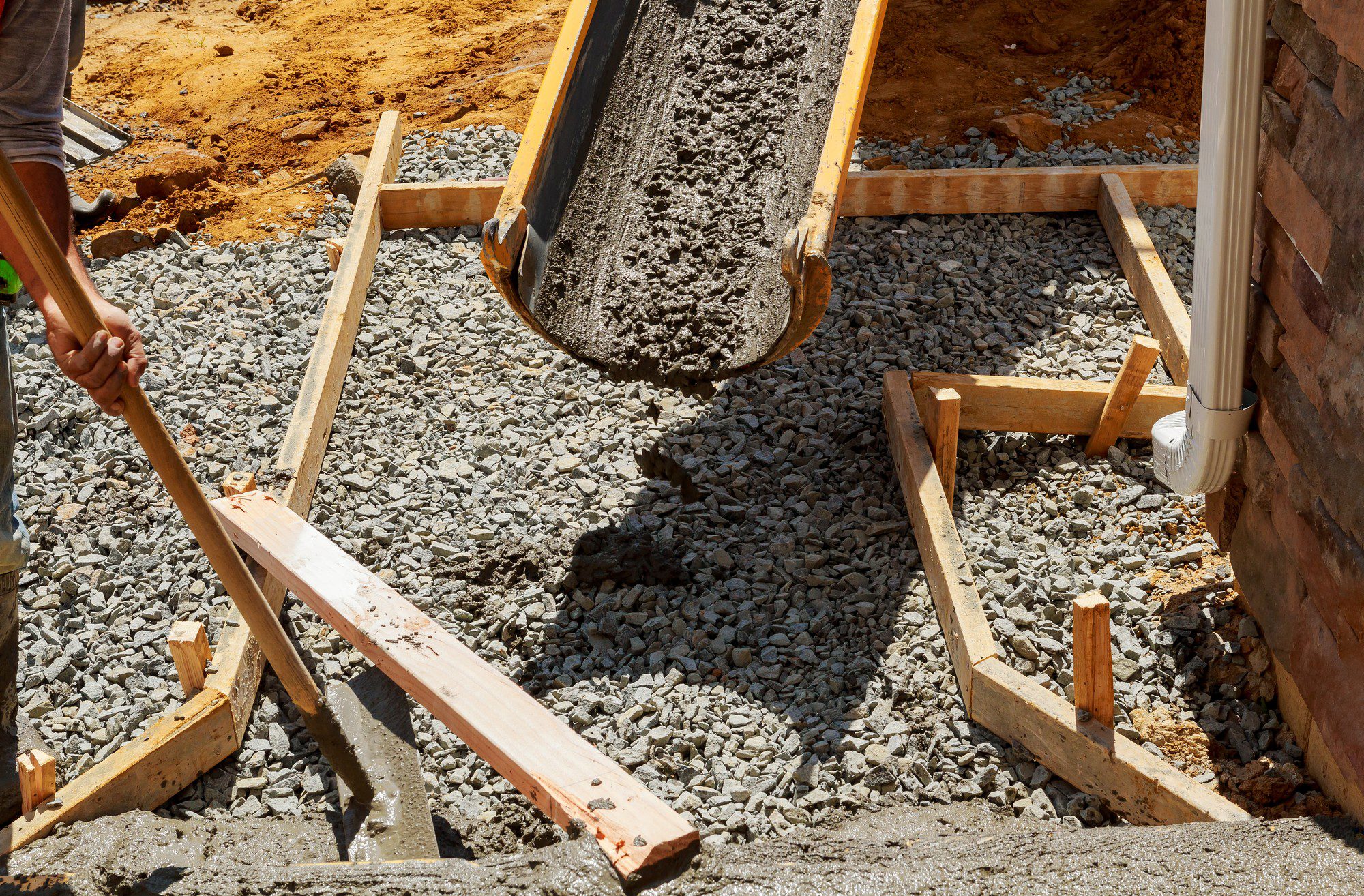 The image shows a construction site where concrete is being poured. The concrete is coming out of a chute, which is likely attached to a concrete mixer lorry, and into a wooden formwork structure. This structure is designed to shape and hold the concrete in place as it cures, and it can be seen filled with gravel, which is often used as a base layer before pouring concrete.There is a person visible, although only partially; the individual is manipulating a shovel, which is usually used to spread and level the concrete. The person appears to be wearing typical protective gear for construction work, like long trousers, and has a visible safety reflector band around their calf, which is often required on construction sites for visibility and safety purposes. The surrounding ground is dirt, indicating that this is an active construction area.