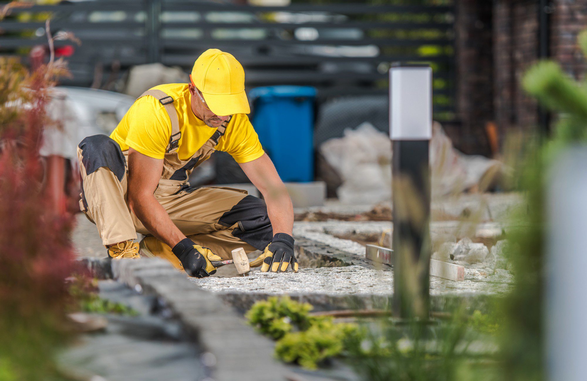 The image shows a person engaged in some sort of construction or landscaping work. The individual is wearing a yellow hard hat and a bright yellow shirt for visibility, which is typical work attire for construction workers. They also have on work gloves and utility pants with knee pads, indicating physical labour. The worker is kneeling down and setting blocks or bricks carefully into place, which suggests they might be working on paving or creating a pathway. In the background, there are some bags that likely contain construction materials and a blue wheelie bin, possibly for waste or additional materials. The focus is on the worker, with plants slightly blurred in the foreground and the background slightly out of focus, creating depth and emphasizing the work in progress.