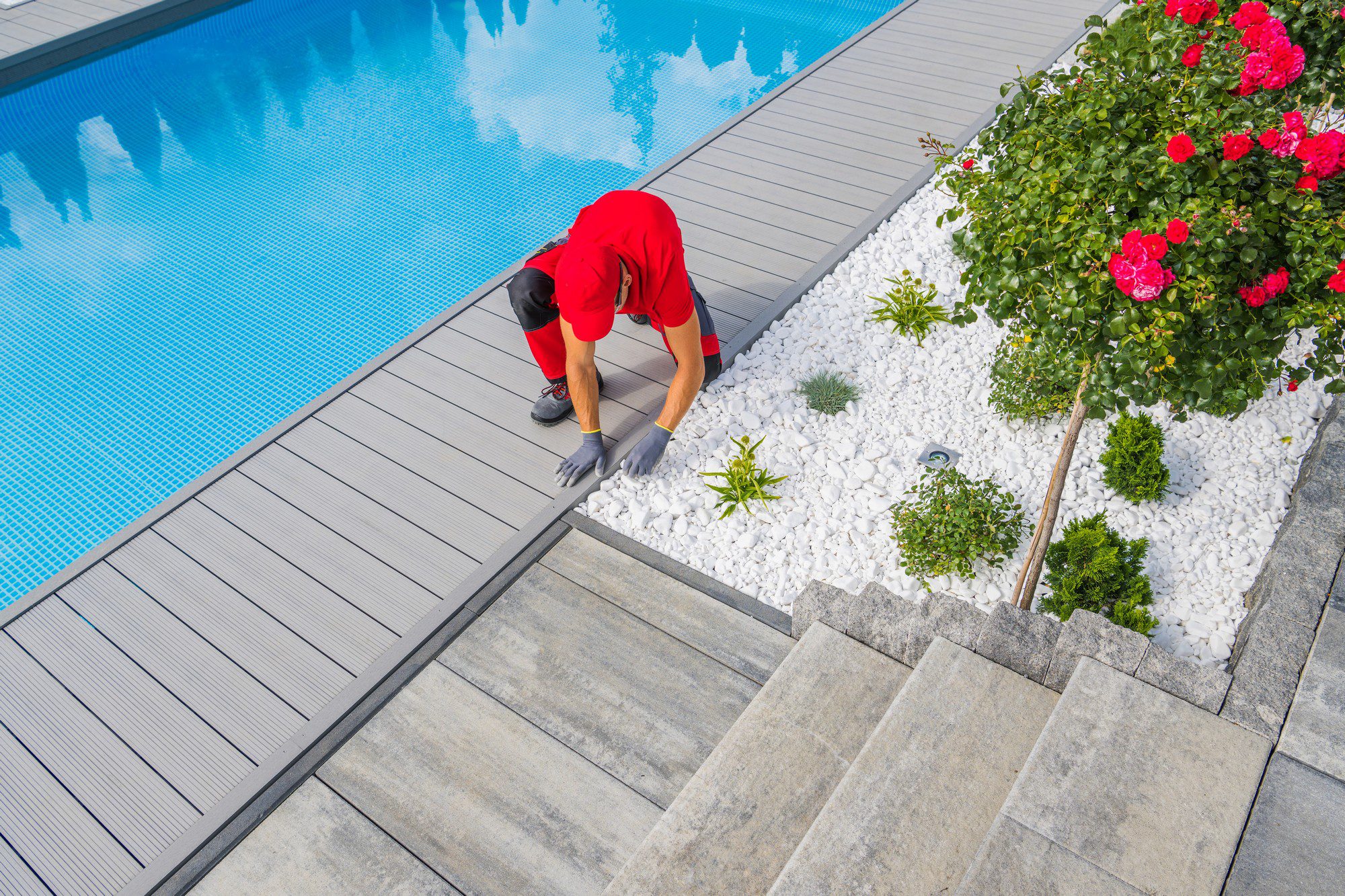 The image depicts an outdoor setting with a swimming pool, landscaping, and a person at work. Specifically:1. A clear, blue swimming pool with calm water reflecting the sky, surrounded by a stylish grey composite decking.
2. A red-shirted individual appears to be performing maintenance or gardening tasks. They are bending over and wearing gardening gloves, suggesting that they might be working with the plants or adjusting the stones.
3. A neatly arranged garden bed with white stones and a variety of green plants, including what looks like a rose bush in full bloom with red flowers, suggesting attentive gardening care.
4. A paved area with large, rectangular stone tiles leading to or from the pool area, contrasting with both the decking and the white stones of the garden bed.
5. The overall setting implies a well-maintained and designed outdoor area, likely in a residential or possibly a commercial location.
The person's presence adds a human element to the scene, indicating care and maintenance of the landscaping and pool area. The image conveys a sense of tranquility with the stillness of the pool and the disciplined design of the garden.